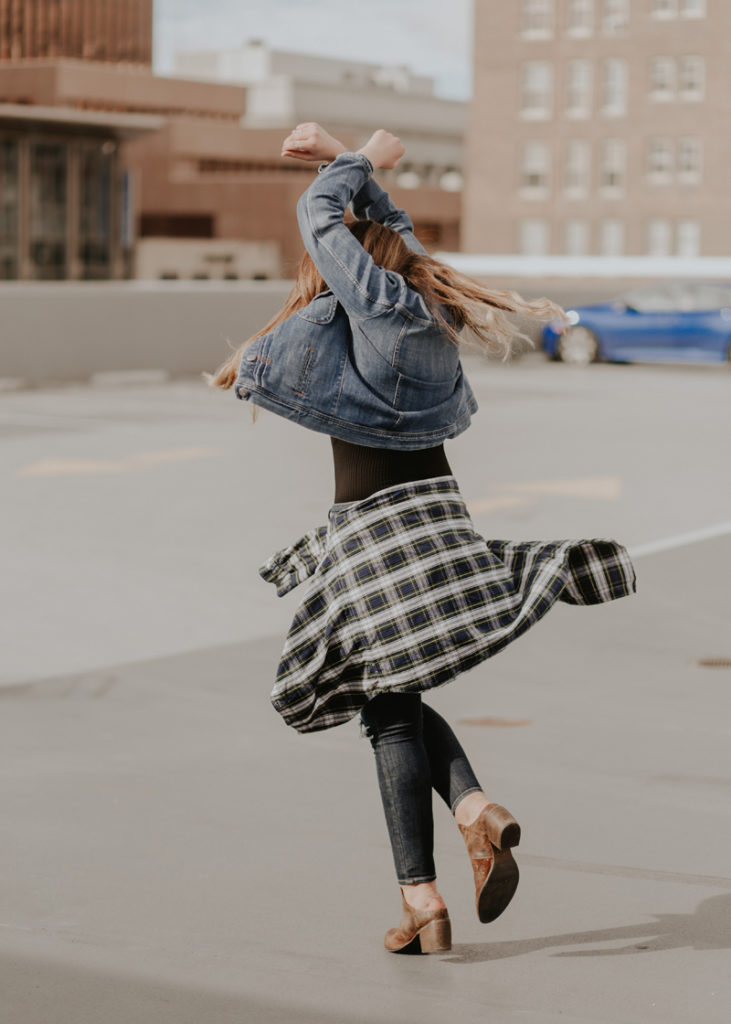Woman wearing jeans and a denim jacket with a plaid shirt tied around her waist spinning and twirling