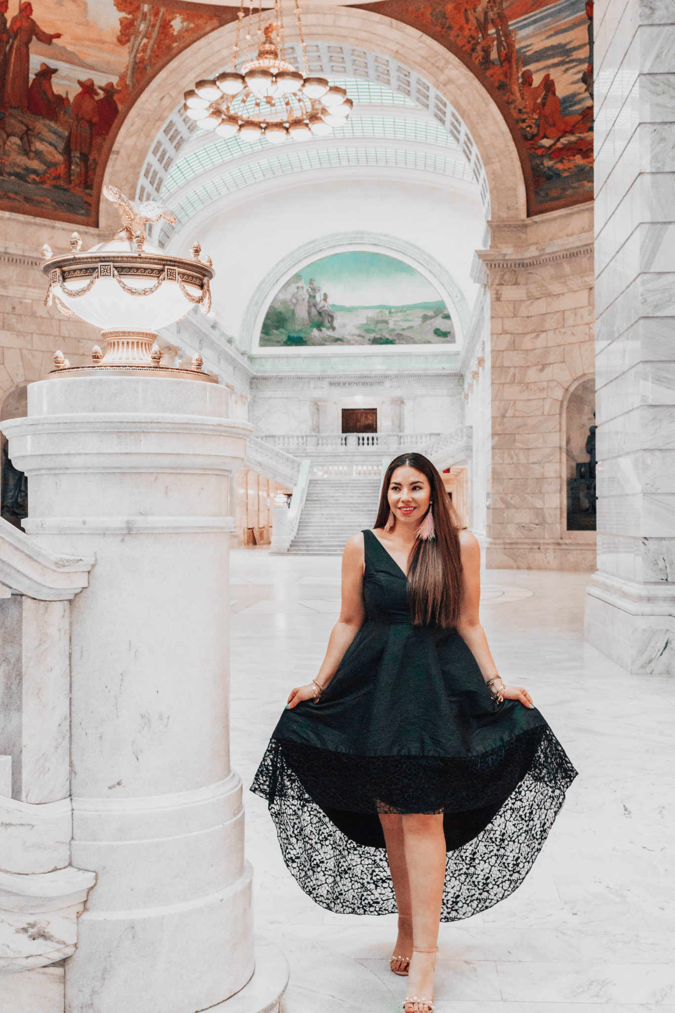 Brunette woman wearing black formal gown and heels walking through large, white marble building.