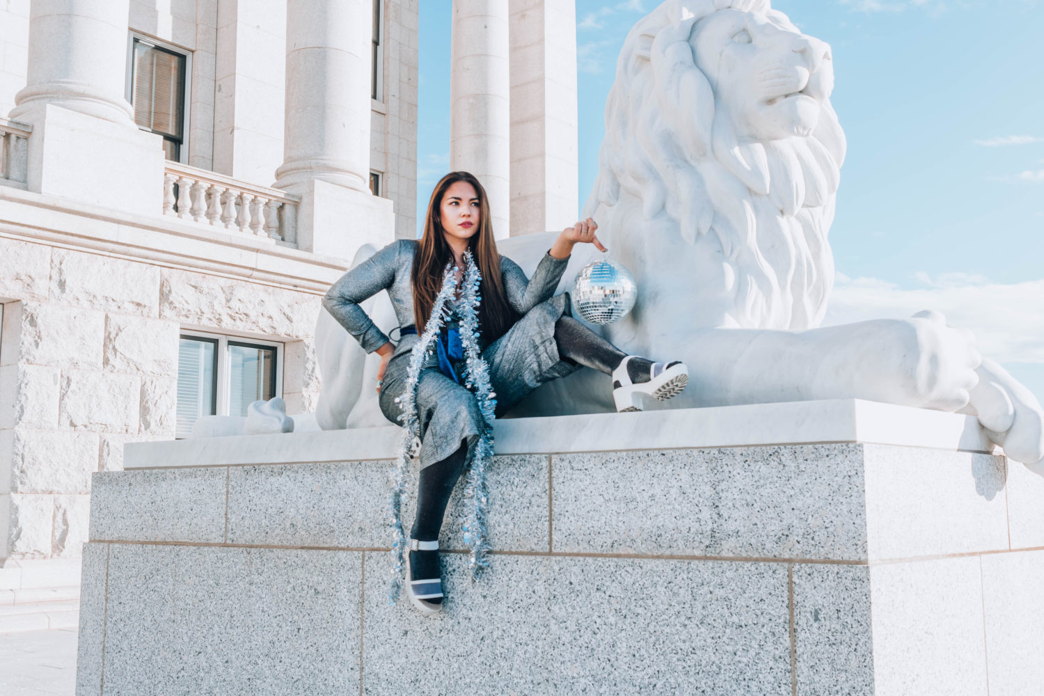 Lauryncakes in grey sequin jumpsuit holding a disco ball and sitting on a sculpture of a lion.