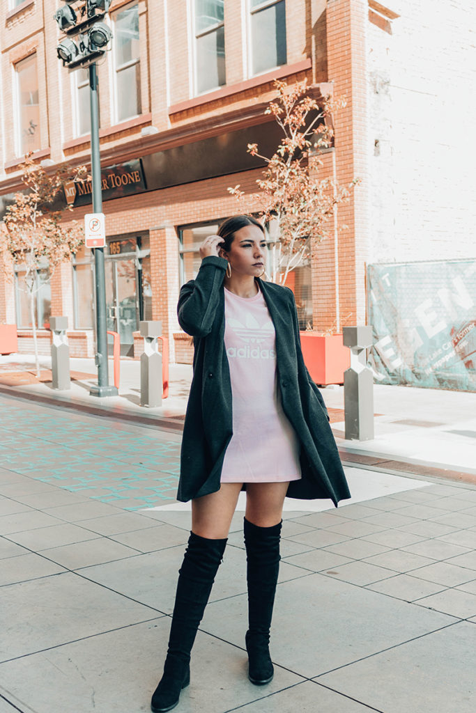 Lauryncakes, fashion blogger, standing in Salt Lake City street wearing tall black boots, a black trench coat, and a pink adidas shirt dress.