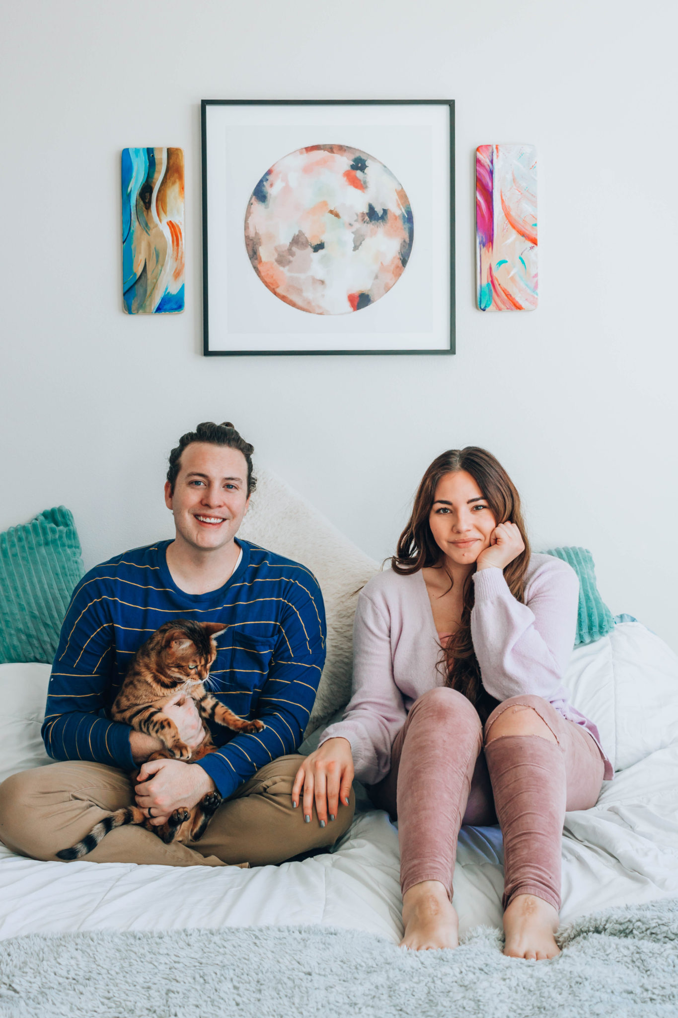 Young couple seated with their cat on their bed beneath their spirit paintings, mounted to the wall behind them.