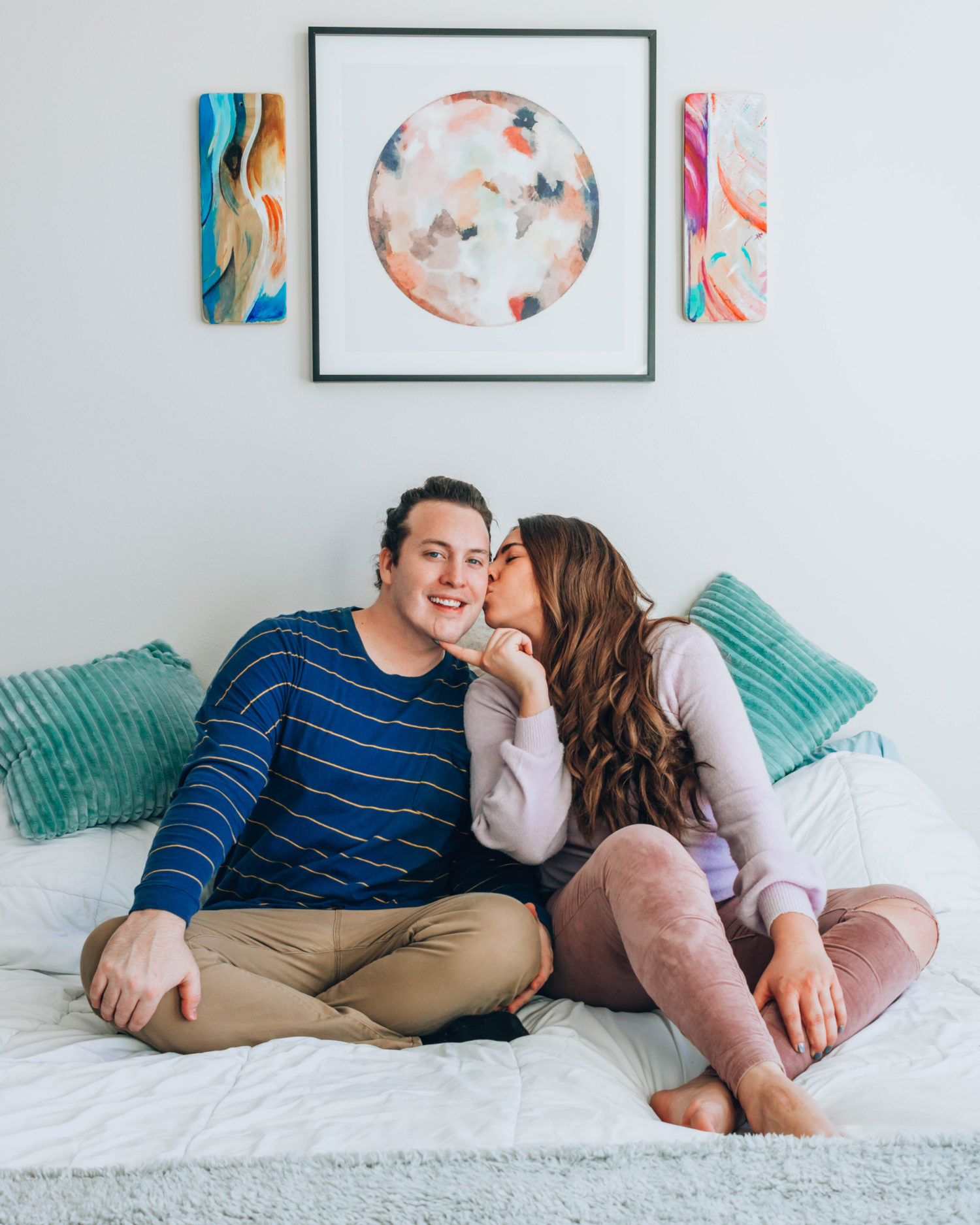 Couple kissing on a bed beneath paintings.