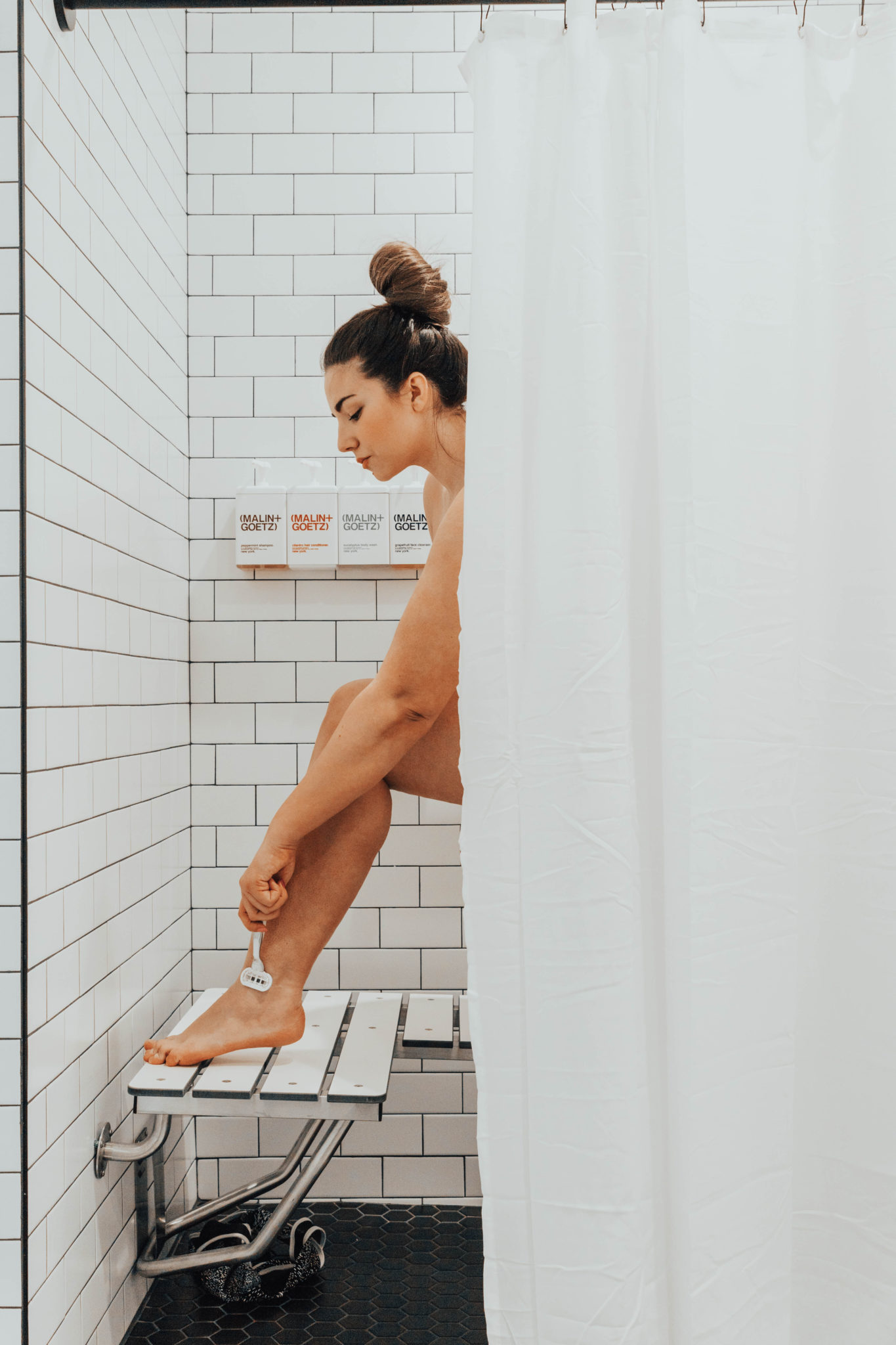 Brunette girl with hair in a bun standing in white tiled shower shaving her legs.