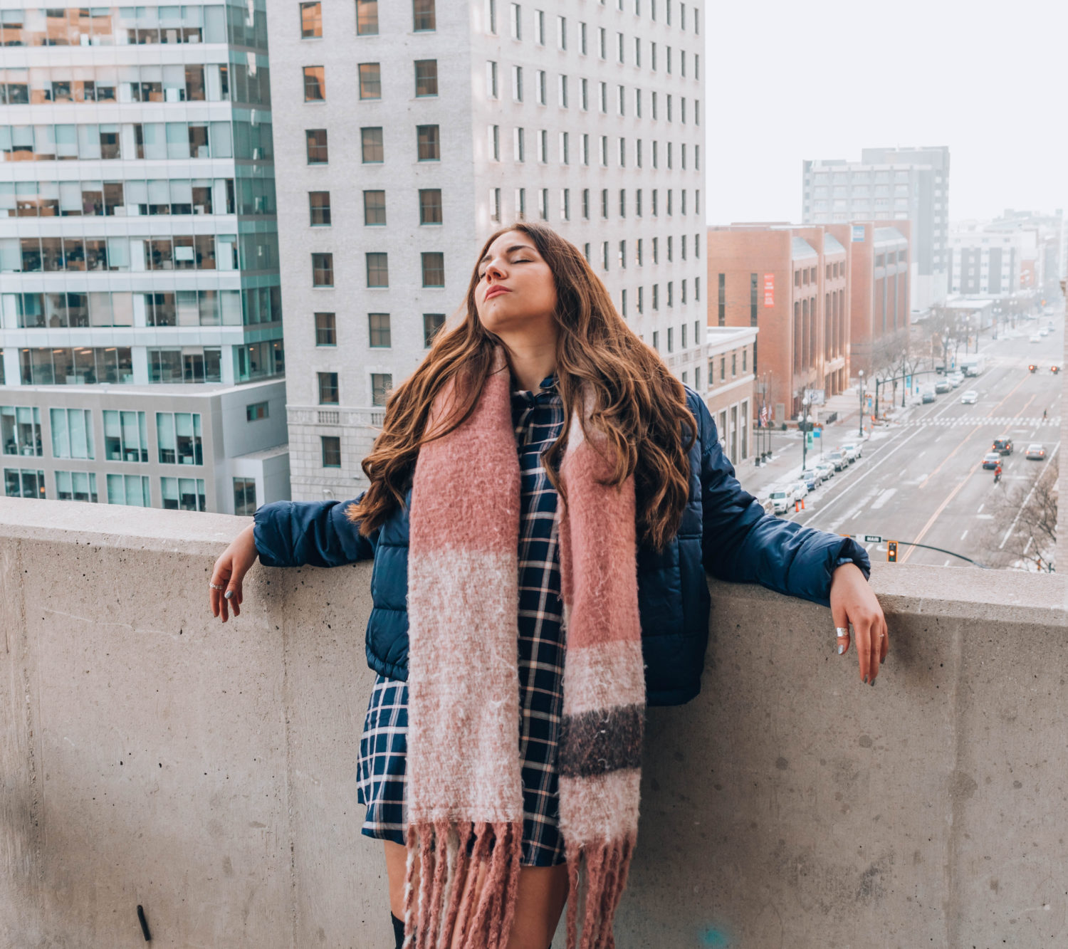 Girl woth long, brunette hair leaning backwards in winter outfit over city buildings.