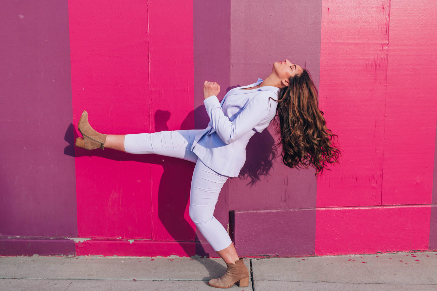 Brunette woman in monochromatic purple pant suit  and tan ankle boots posing  with leg up in the air. 