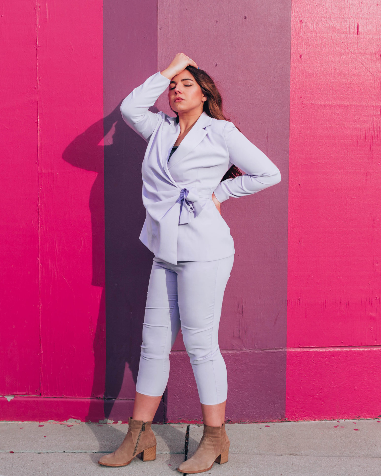 Woman in monochromatic purple pant suit posing dramatically in front of a pink and purple striped wall.