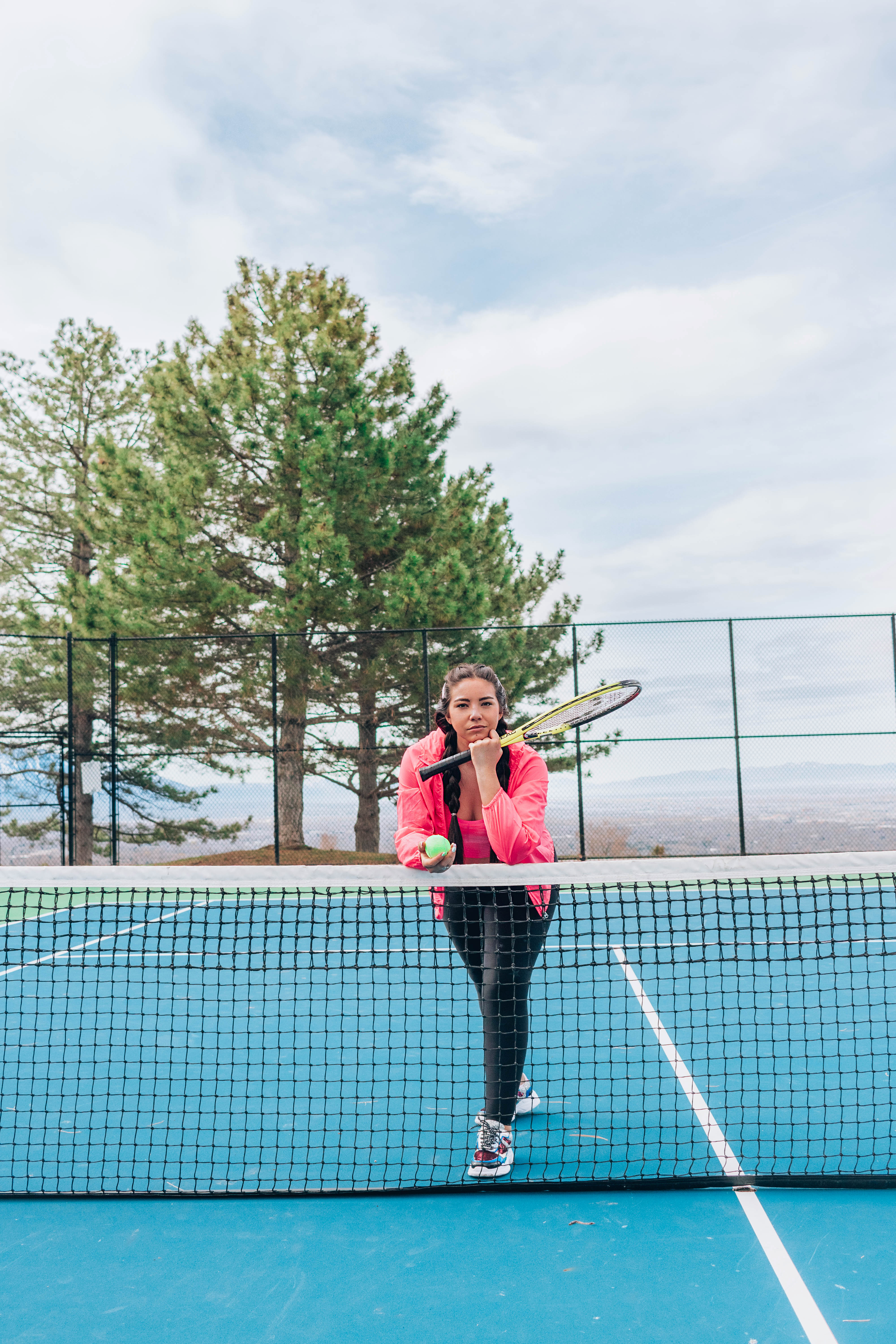 Woman wearing a pink jacket and workout clothing leaning on a tennis net holding a ball