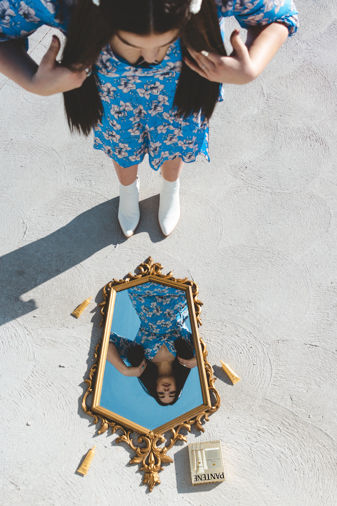 Lauryncakes in a long floral, blue dress and white boots looking into her reflection of a large gold mirror placed on the ground
