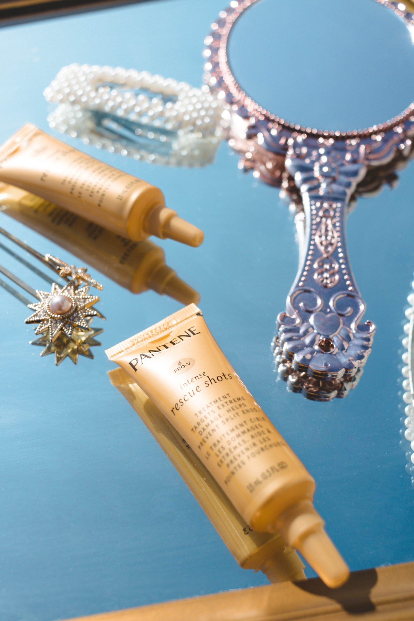 Close-up shot of golden Pantene Pro-V Intense Rescue Shots sitting on a reflective surface with a handheld mirror and barrettes laying next to it.