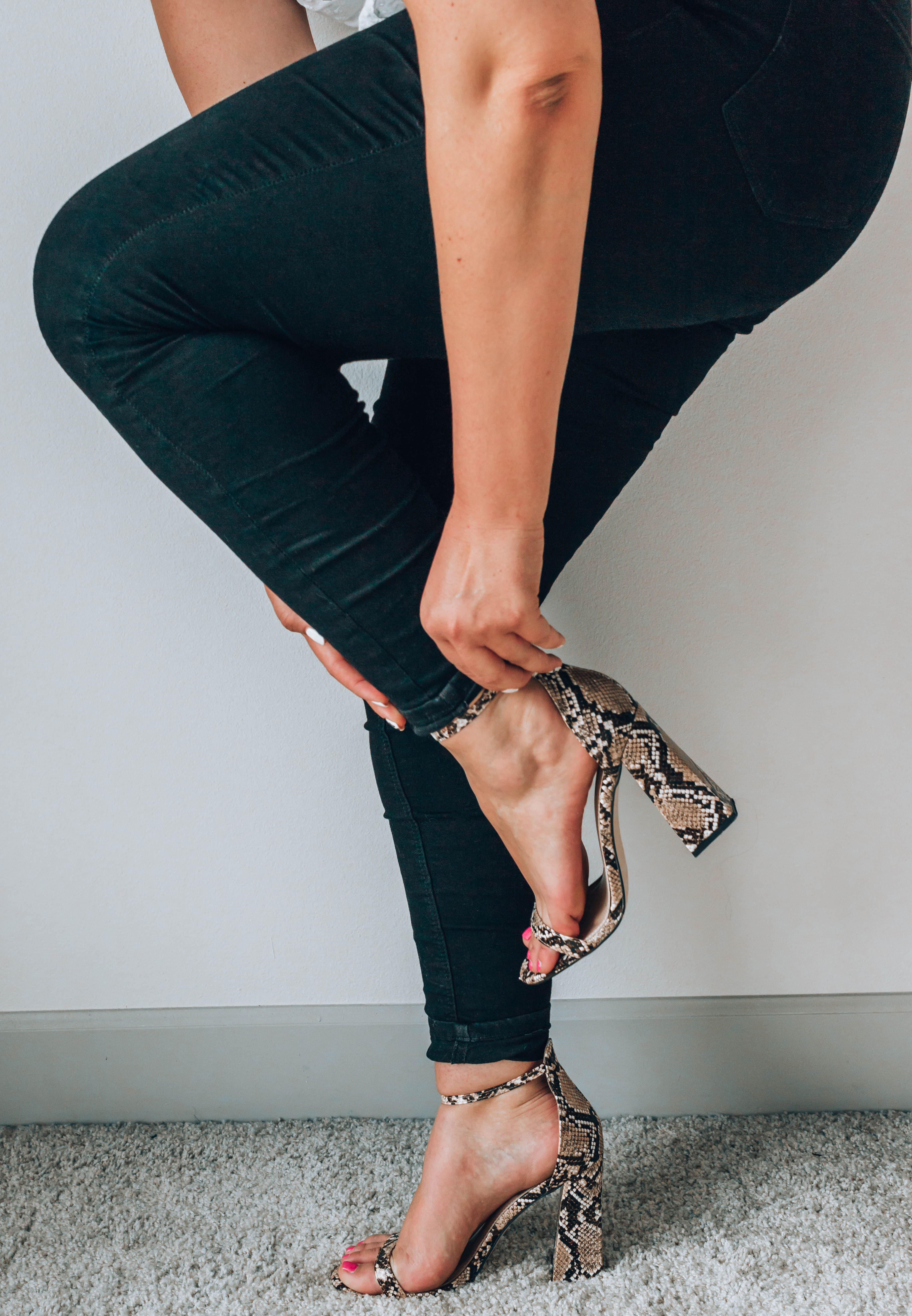 Photo of a woman wearing black skinny jeans holding her ankle strap snakeskin heels