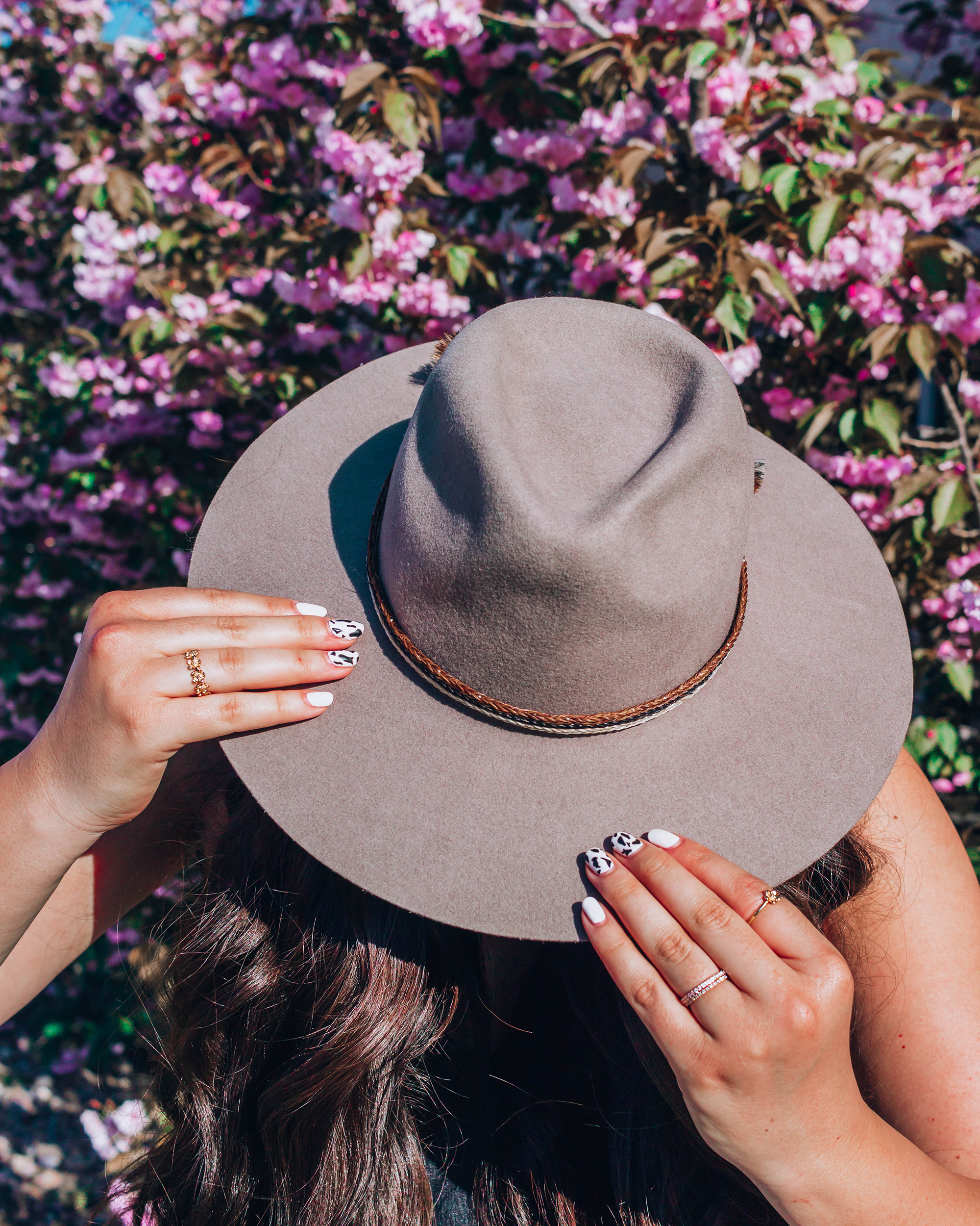 Boho, western style wide brim hat being held in hands with gold jewelry on them in front of a floral background