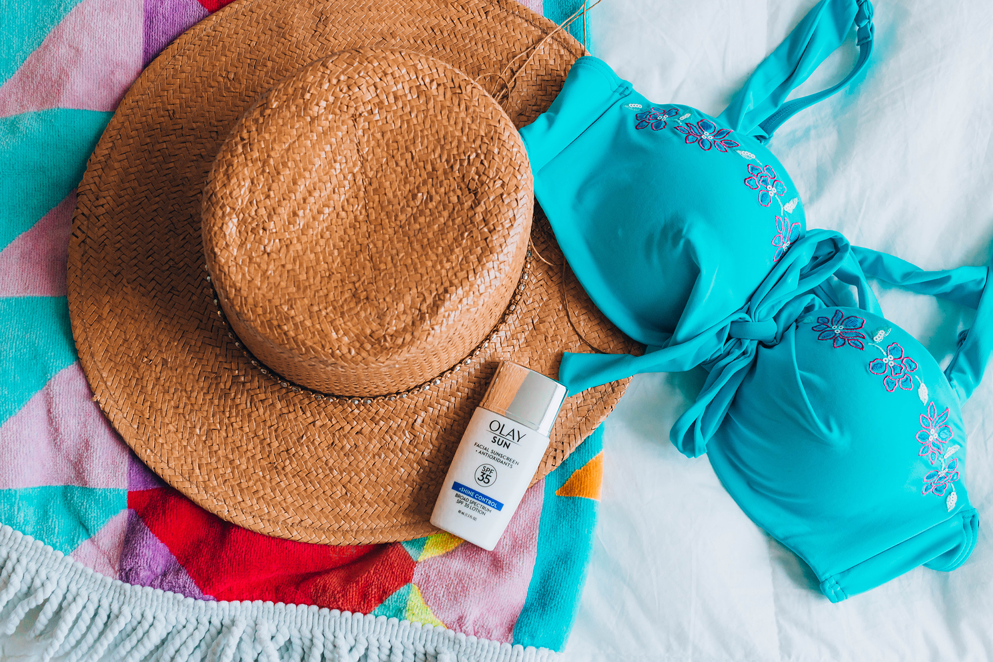 beach accessories (including a straw hat, bikini top, rainbow towel, and Olay SPF moisturizer) in a flatlay 
