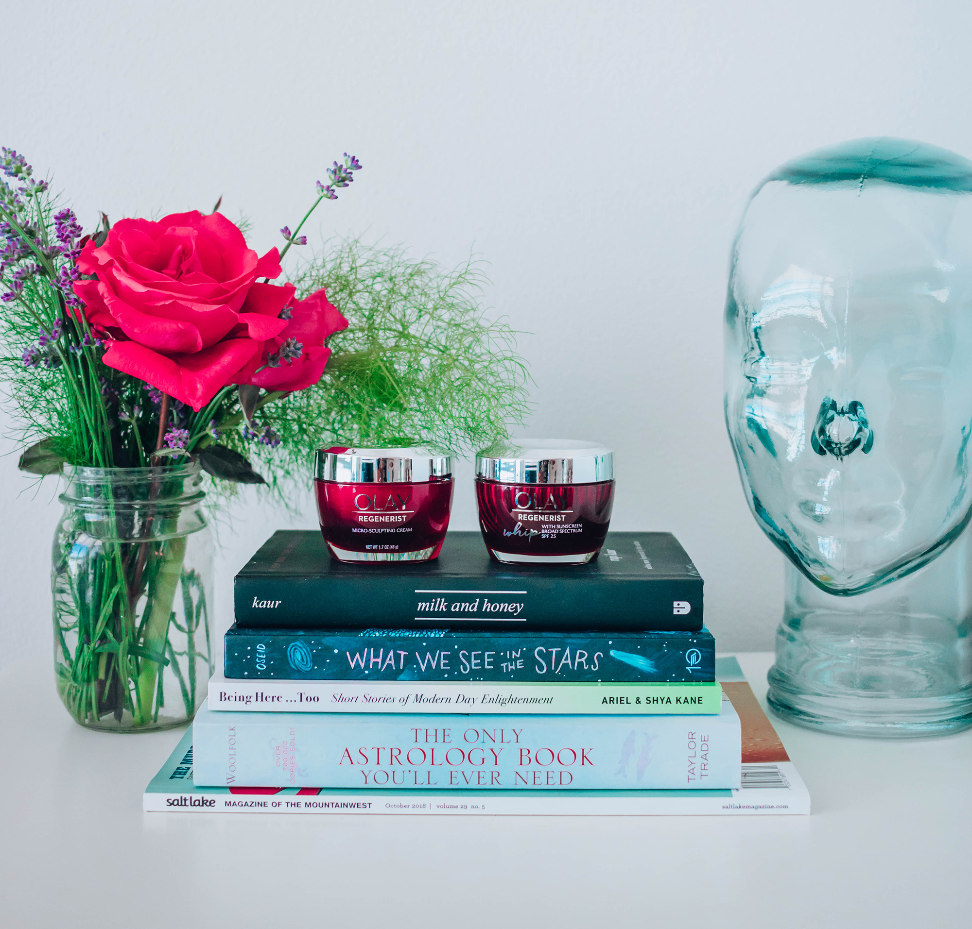 Olay Micro-sculpting Cream and Olay Whip read jars sitting on top of stacked books next to a rose bouquet and glass head art