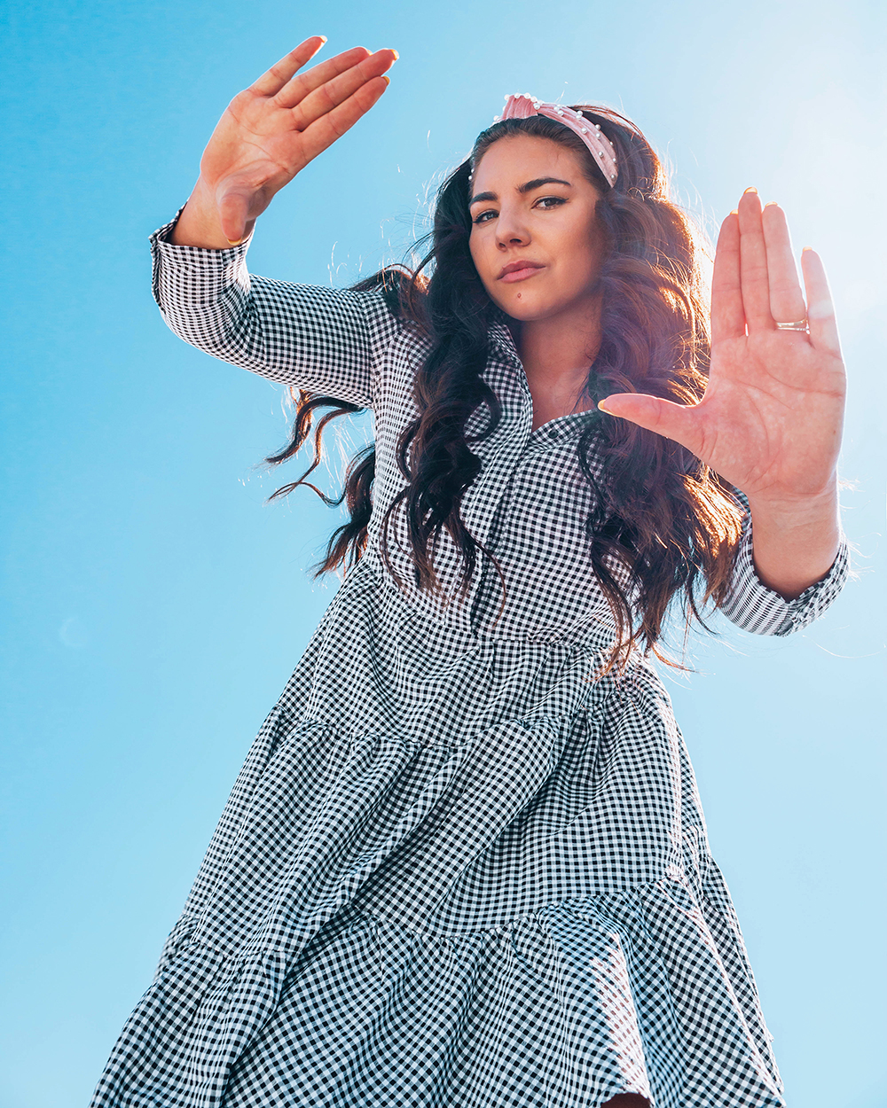 Lauryn Hock facing the camera and holding her hands up to frame it. She's wearing a gingham dress and big, pink headband on her loosely curled hair