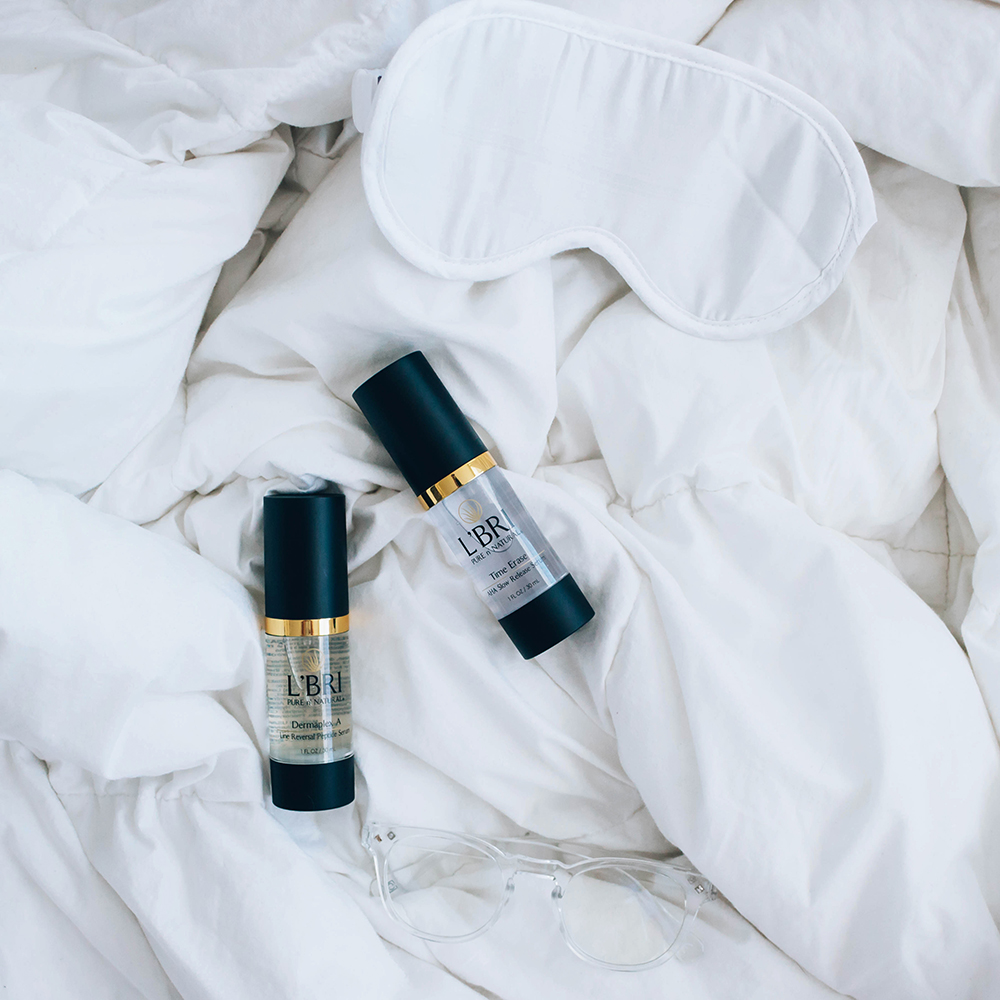 Skincare serums in a flat lay on white sheets next to an eye sleep mask and reading glasses