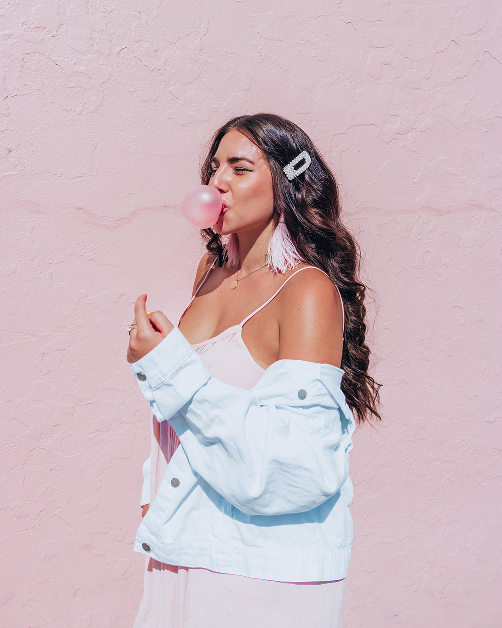 Lauryn Hock standing sideways in front of a pink wall. She's closing her eyes and blowing a bubble while wearina pink dress, white denim jacker, feather earrings, and a pearl hair clip.