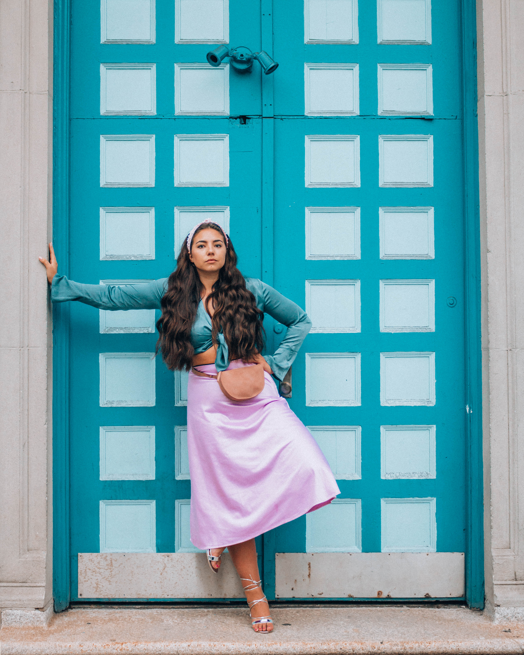 Lauryn Hock from Lauryncakes standing outside of a blue door wearing a blue silk top and a pink silk skirt while her long curled hair is pulled back with a head band