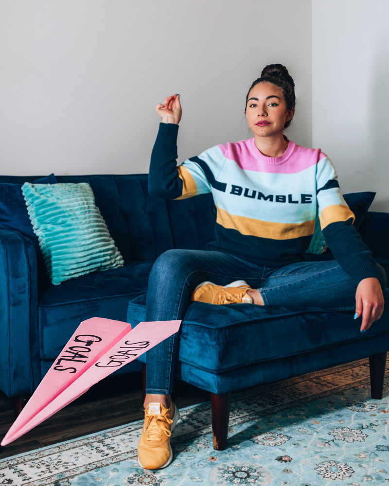 Lauryncakes sits on a blue bvelvet couch while wearing a rainbow striped sweater skinny jeans, and sneakers. She's throwing a paper airplane that has "goals" written on it