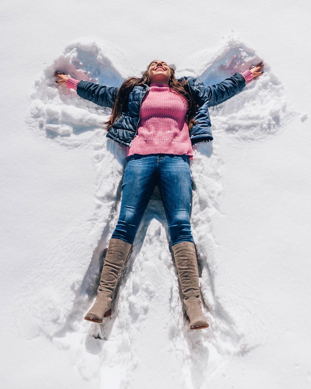 Lauryn Hock making a snow angel while wearing a winter outfit of tall boots, jeans, a sweater, and jacket