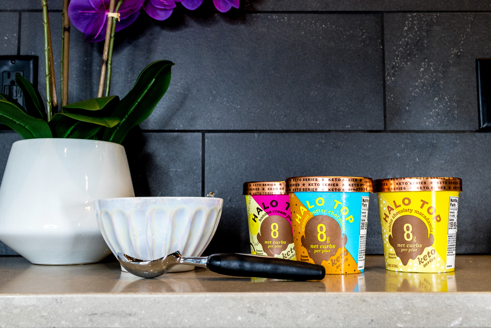 Halo Top® Keto Series Pints photo showing Halo Top Keto White Chocolate Macadamia, Halo Top Keto Peanut Butter Chocolate, Halo Top Banana Cream Pie on a counter next to an ice cream scoop, bowl, and pink flowering plant