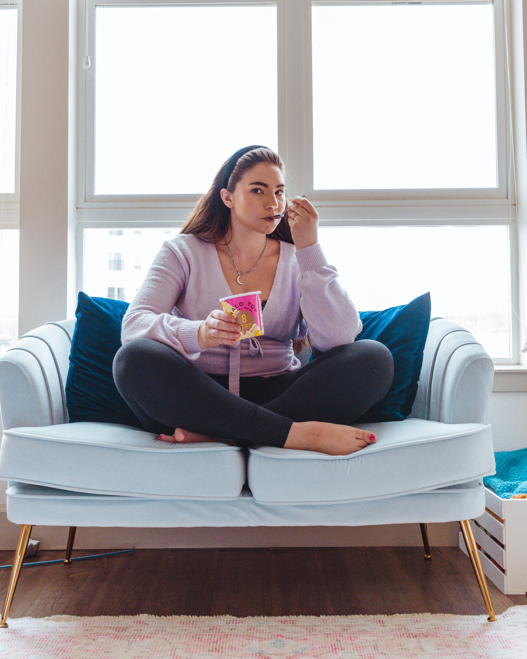Lauryn Hock (blogger at Lauryncakes) sits on a blue velvet couch wearing loungwear and eating Halo Top Banana Cream Pie Keto