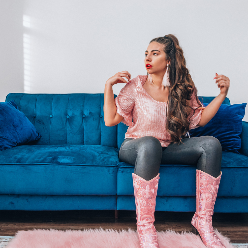 Lauryn Hock sits on a blue velvet couch in Downtown Salt Lake City while wearing a sparkly top and boots and Spanx leggings