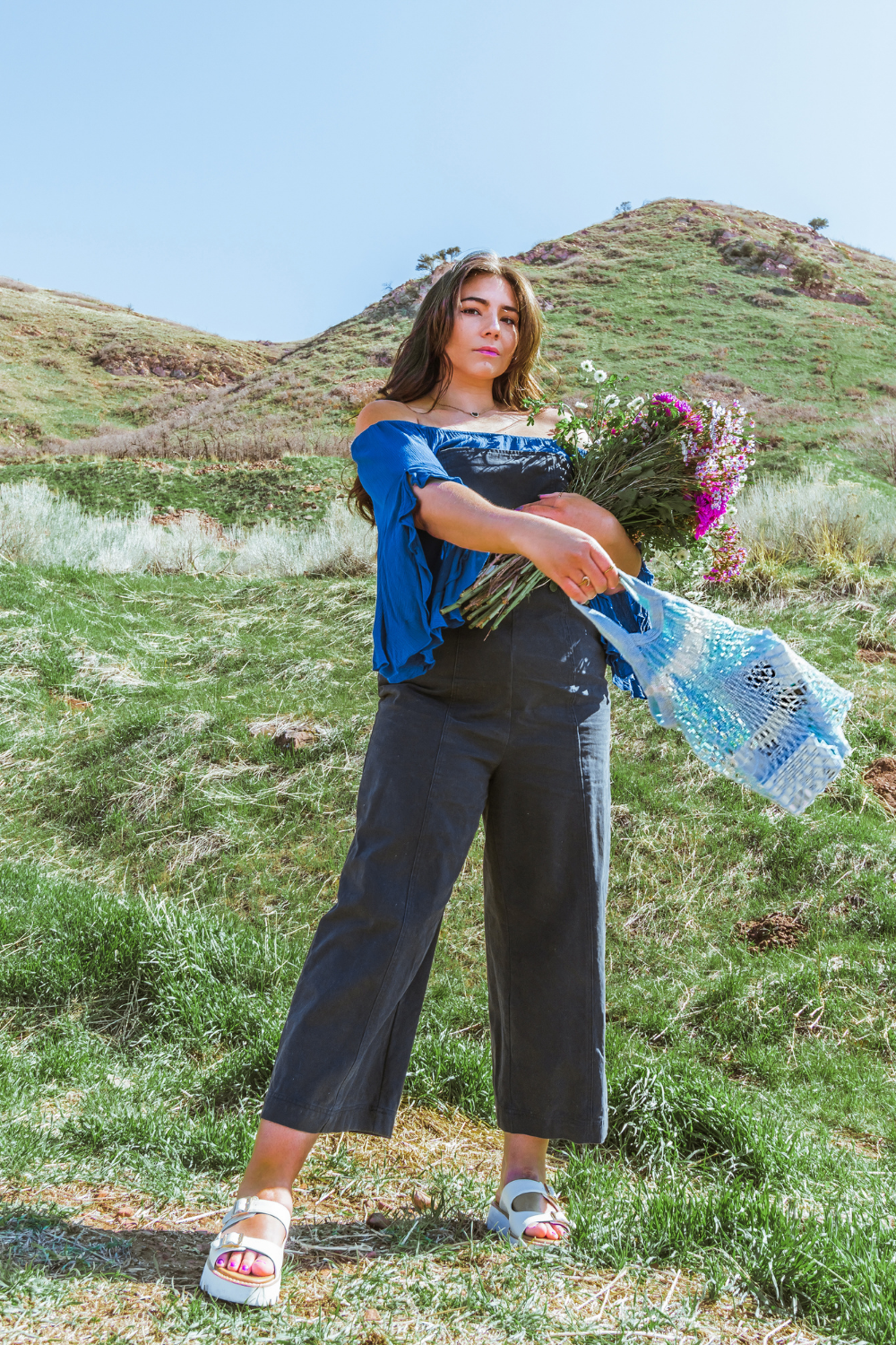 Midsize fashion blogger Lauryn Hock of Lauryncakes social media stands in a green field of grass near Downtown Salt Lake City holding a bouquet of flowers and wearing blue overalls and an off the shoulder top. She has a netted reusable bag in her hand