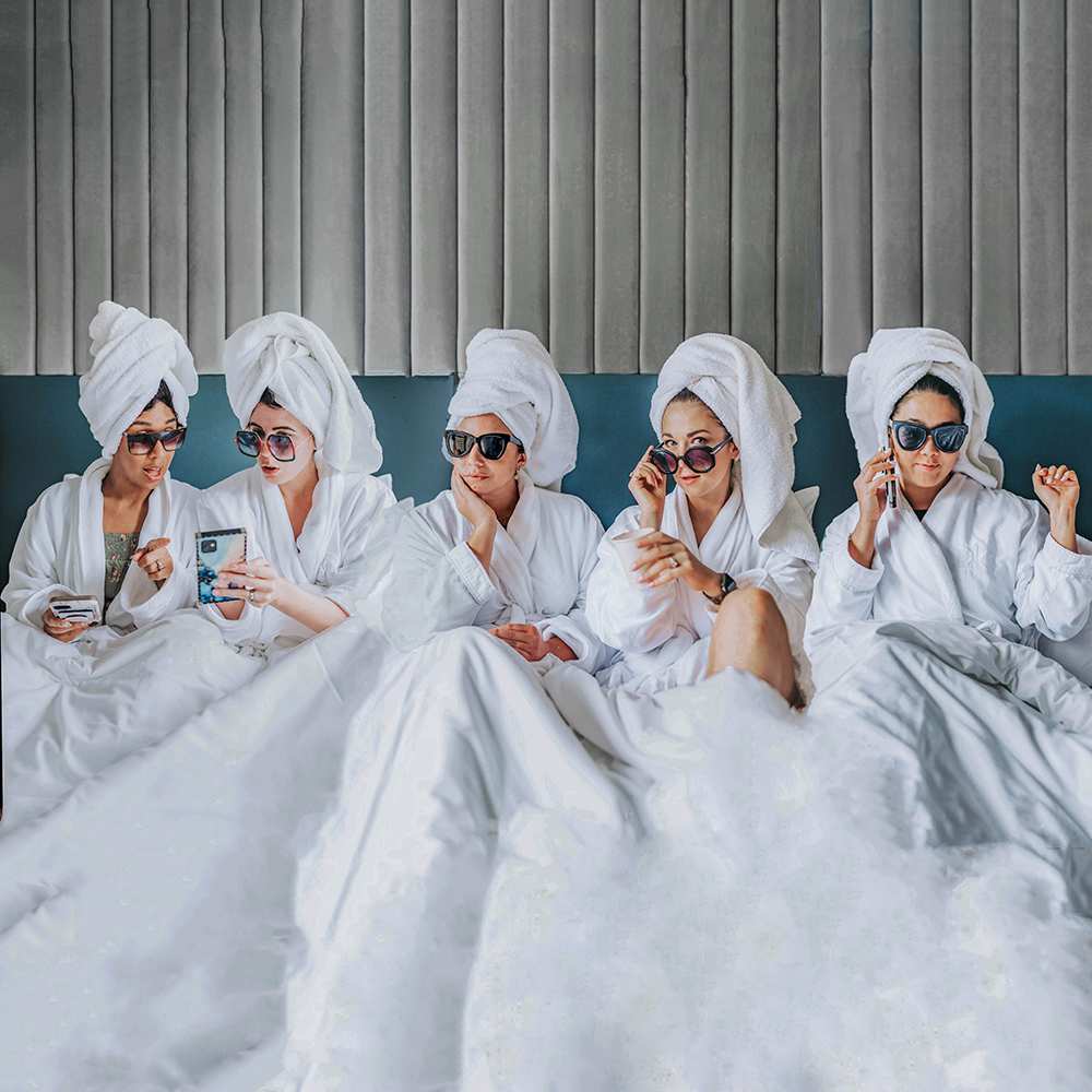 five women with towels on their heads and wearing robes sit under a blanket in a hotel bed. They are on their phones and wearing sunglasses while they lounge