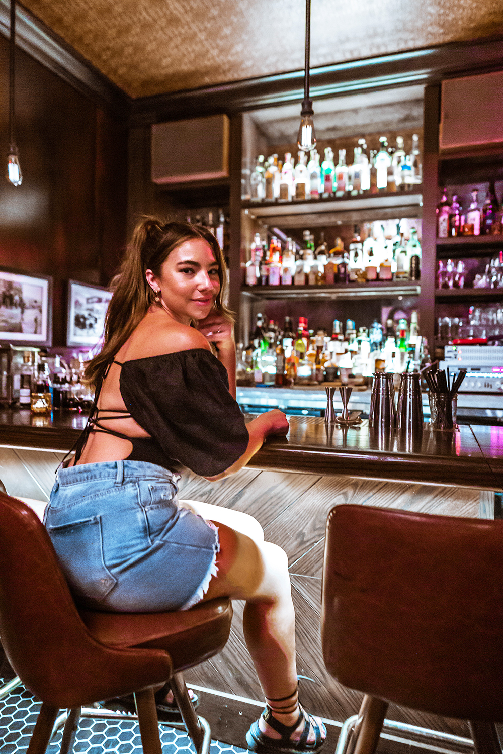 A woman (Lauryncakes) turns around on a barstool. She's wearing a backless bodysuit, denim shorts, and lace-up sandals with her hair in a ponytail