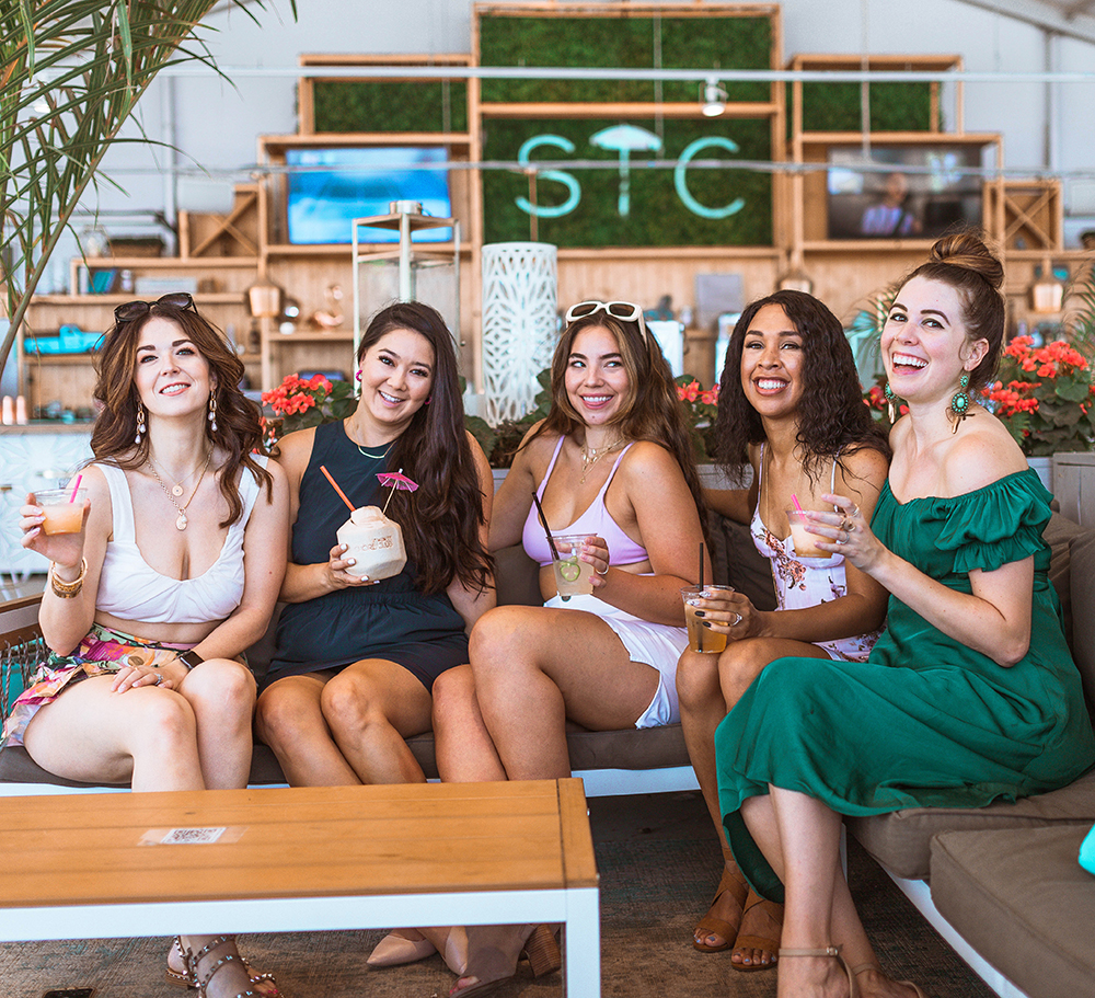 five women in beach clothing sit on a couch holding cocktails at The Shore Club a Chicago Restaurant
