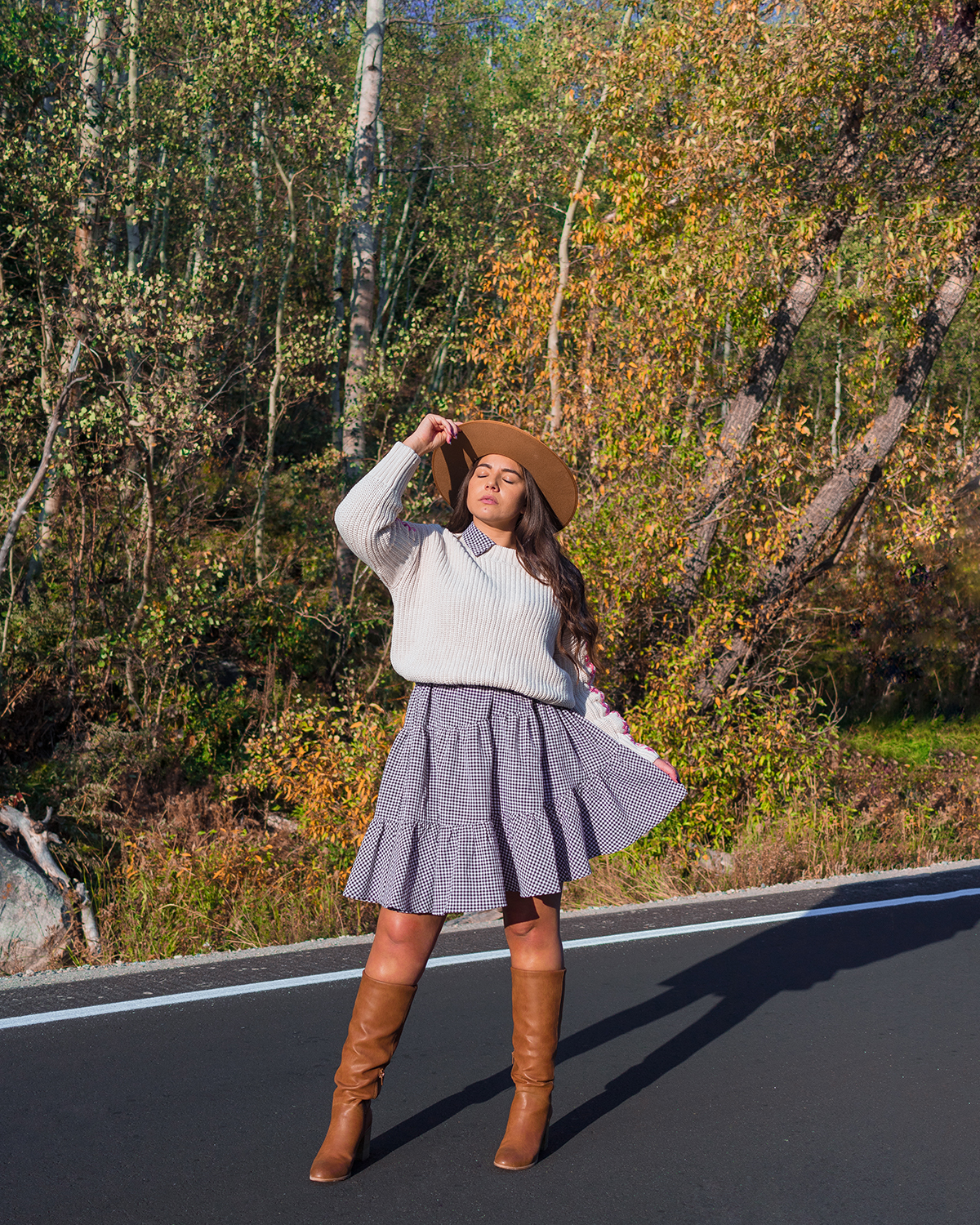 Lauryn Hock in Cottonwood Canyon wearinga Fall outfit of a dress layered under a sweater with knee-high boots and a wide brim fedora. Behind her are fall leavees