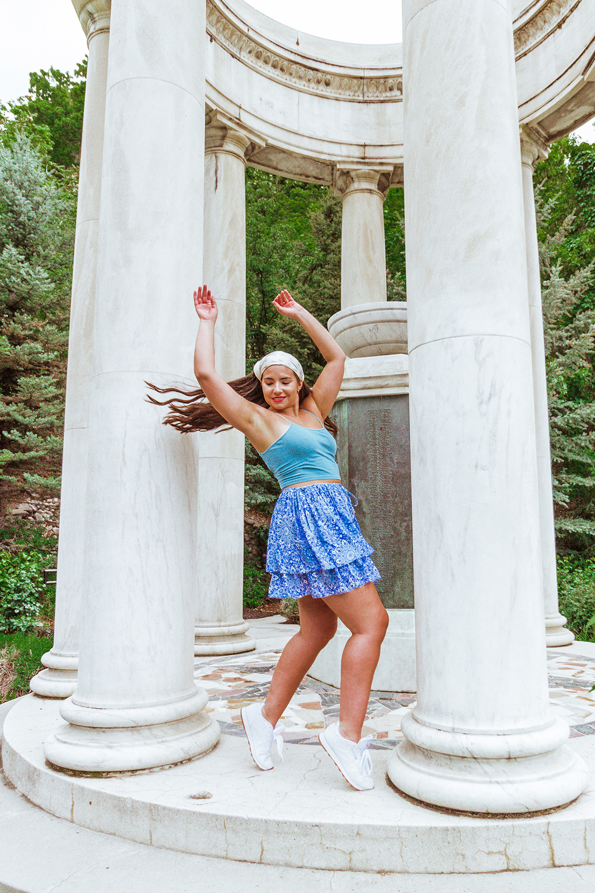 a woman stands on her tip toes spinning under tall marble columns in a park. Her corse, freshly washed hair is in the air behind her while she wears sneakers, a mini skirt, a tank top, and a bandana on her head