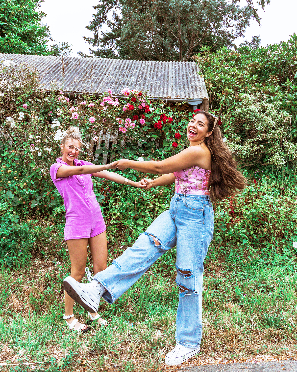 two women hold both hands and look like they are spinning and laughing wearing summer outfits in front of green shrubbery and blooming Oregon flowers. The brunette's hair flies behind her