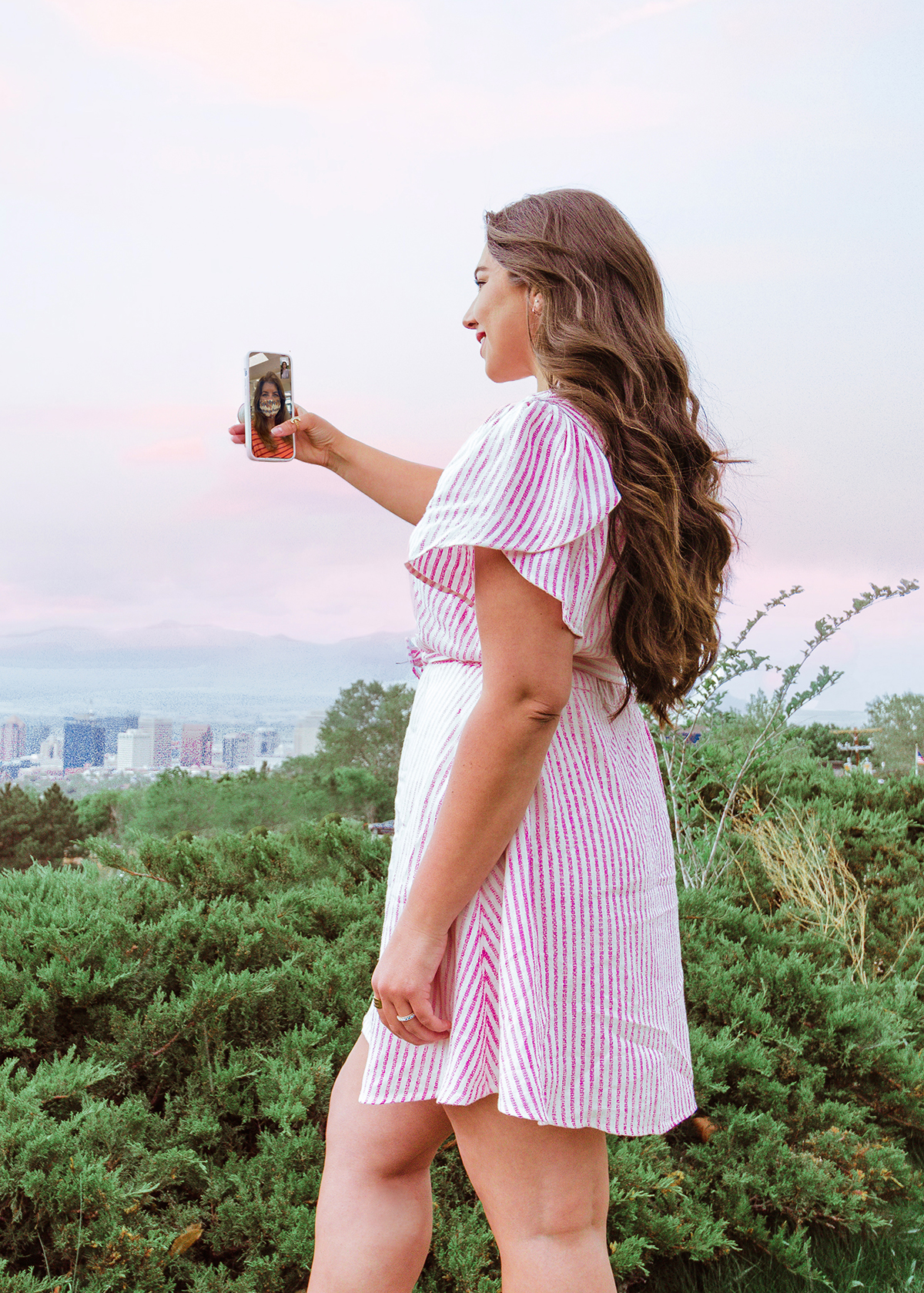 woman facetimeing on the phone with an overlook over Downtown Salt Lake City, Lauryncakes is wearing a pink striped dress with her shampooed tresses curled while she stands in front of bushes