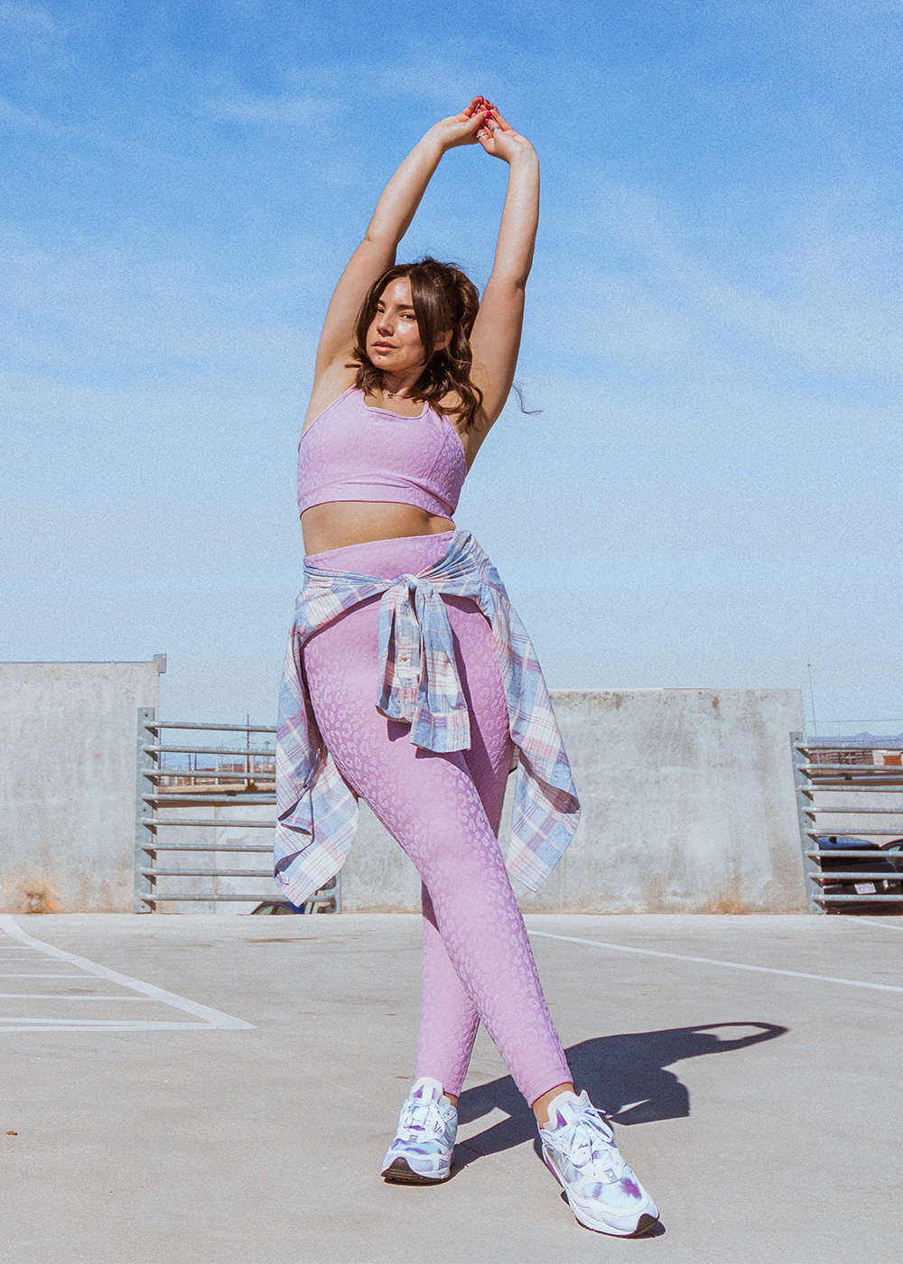 woman poses with her hands over her head wearing a pastel colored barre class workout set of leggings and sports bra with leopard print embossment. around her waist a plaid button up shirt is tied and her hair is pulled back in a ponytail with bangs framing her face. The sky is blue behind her and she's standing on the roof of a parking garage