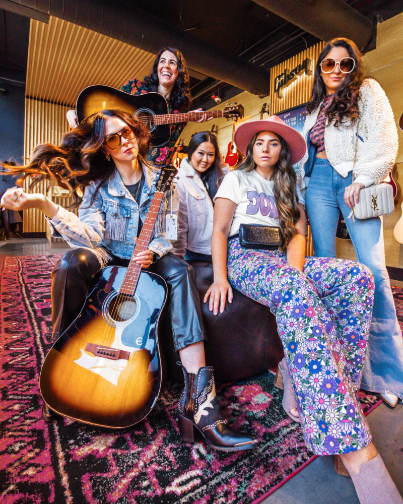 a group of five woman (Babes That Wander)dressed in rock and roll seventies clothing stand and sit next to each other while holding two guitars.