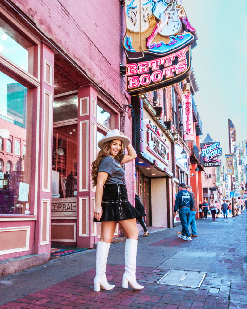 Fashion, beauty, and travel blogger Lauryn Hock wearing a western style outfti of a wide brim fedora hat, band tee, suede skirt, and white knee-high boots. She's walking on Broadway street in Nashville
