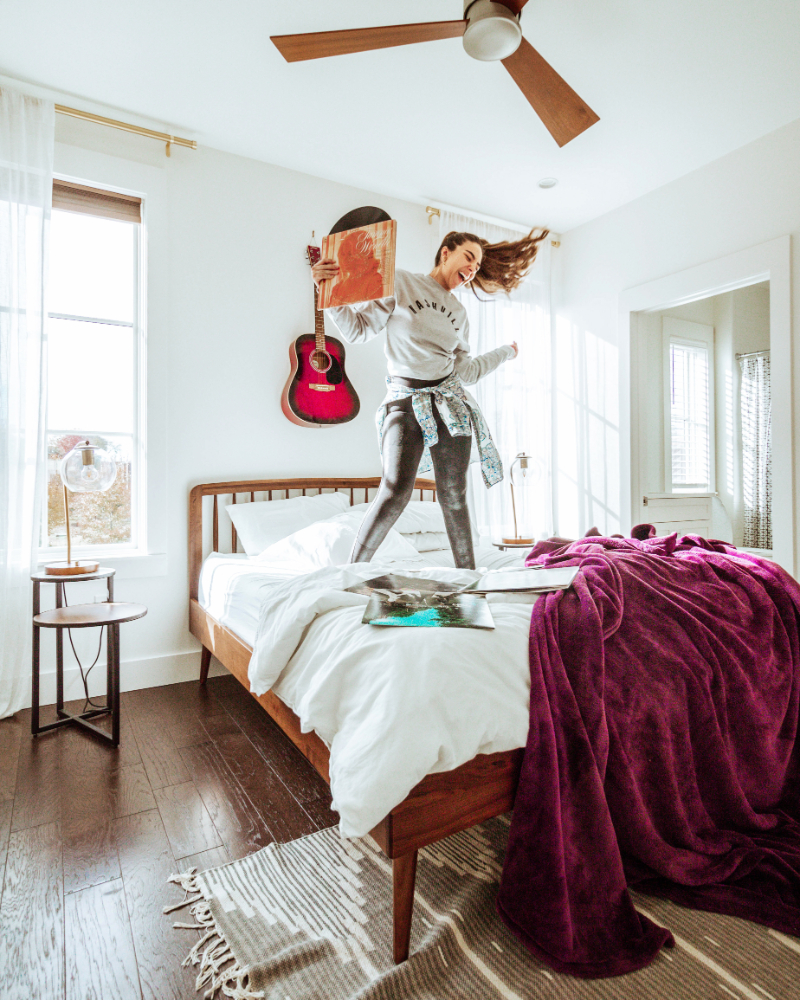 Woman dancing and singing while flipping her hair and standing on a bed. She's holding a record and a guitar is behind her on the wall