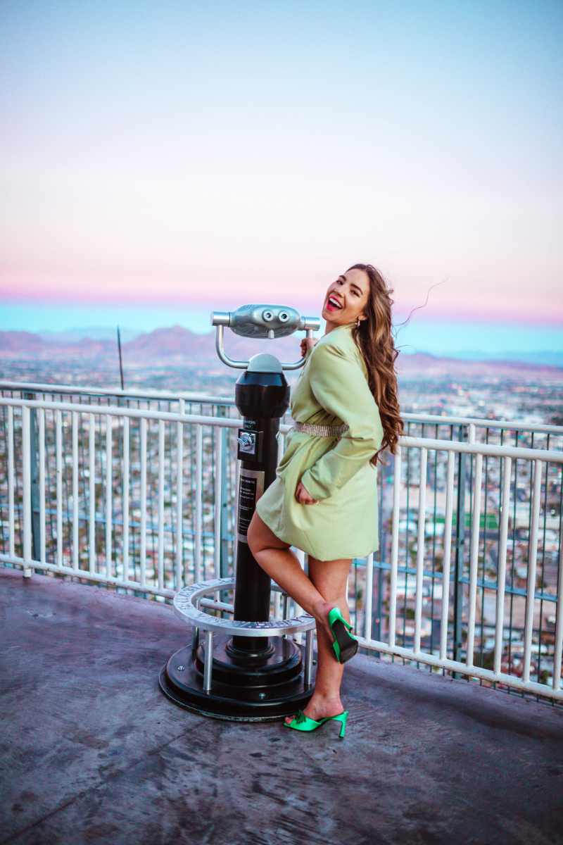 Lauryn Hock laughs and poses in green heels and an oversized blazer. The travel blogger stands near observation binoculars at The Skypod at The Strat during sunset