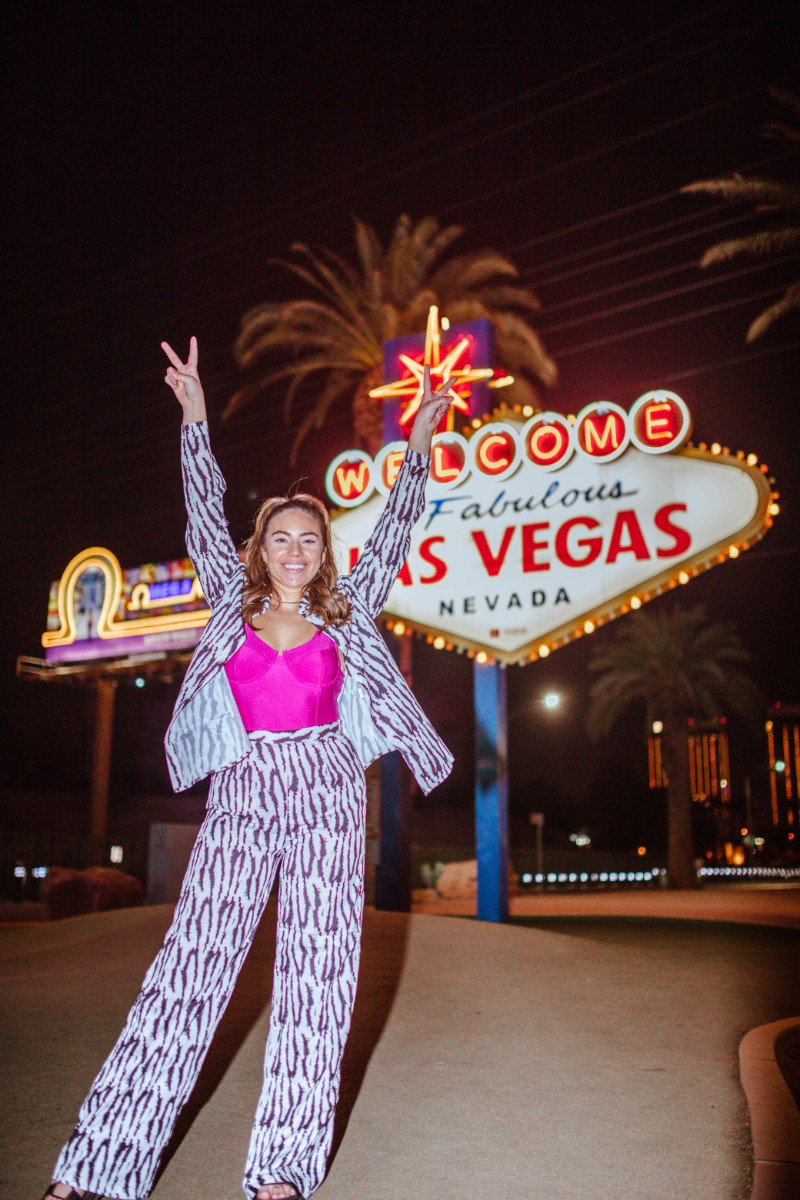 Old Las Vegas Sign to see for tourists. Woman wearing a zebra print pants and blouse set does peace fingers while standing in front of the neon sign at night