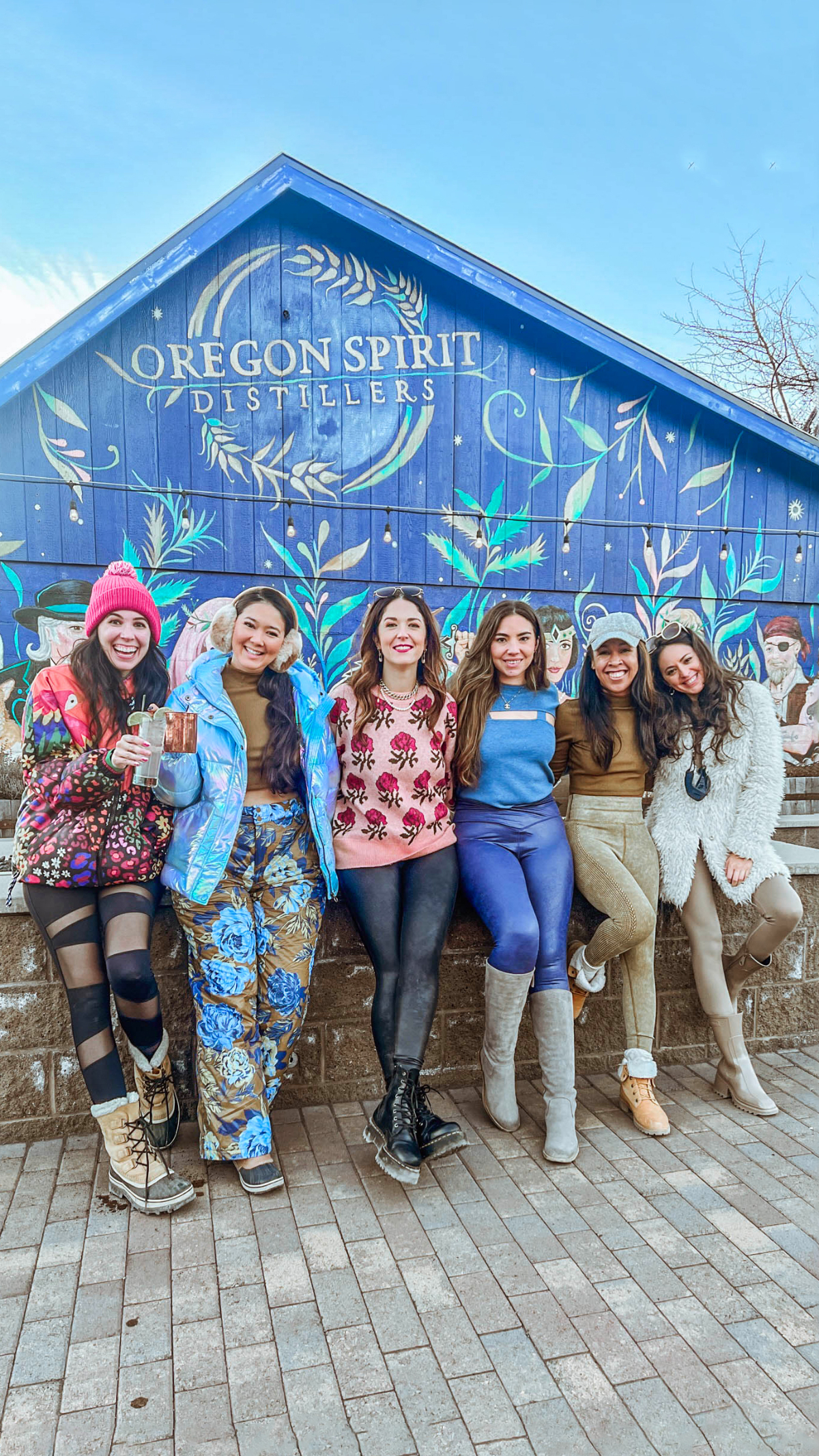 The women of Babes That Wander stand side by side smiling while standing in front of a mural at Oregon Spirit Distilleries