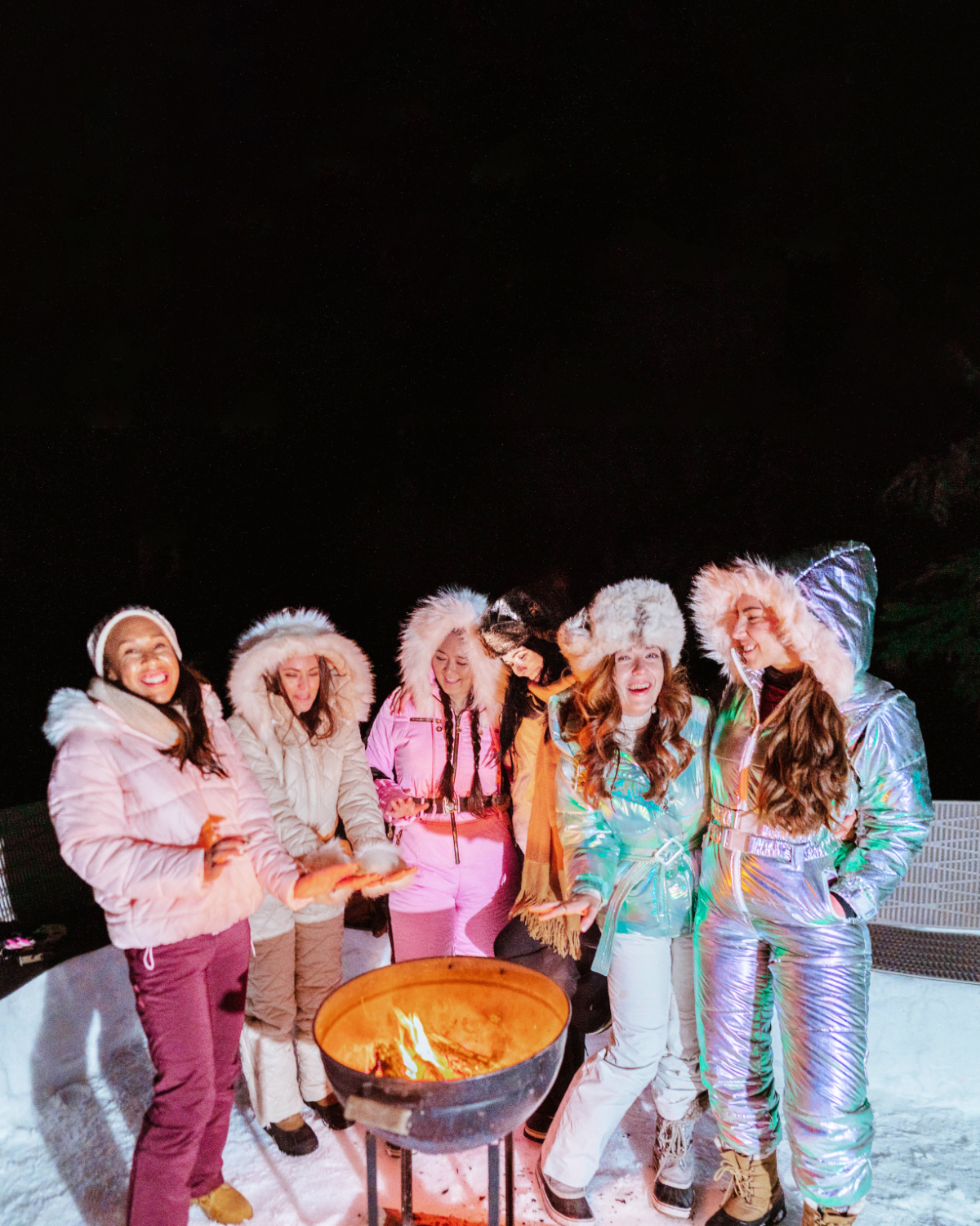 Babes That Wander women travel group stand around a fire talking and laughing after snowshoeing at night