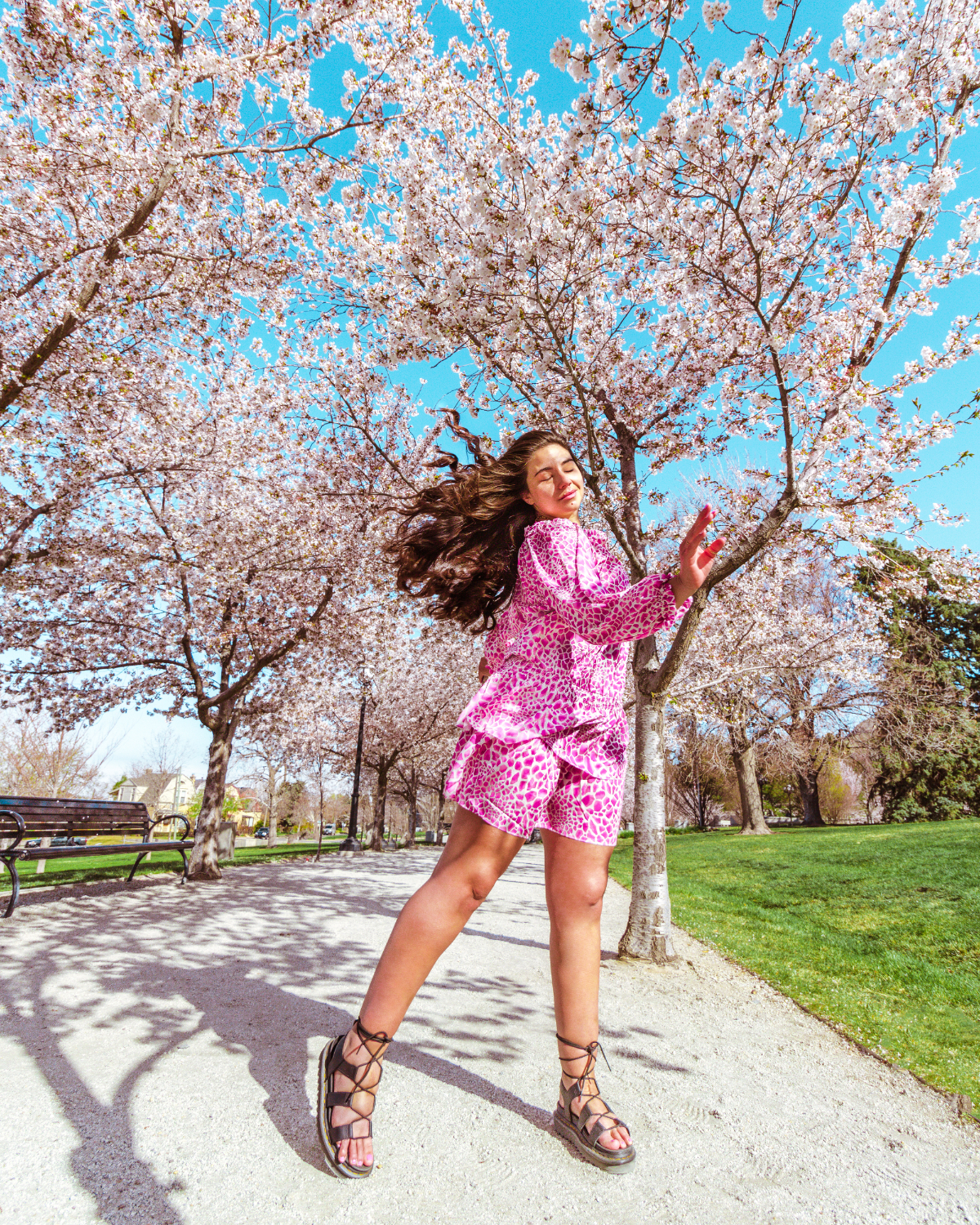Utah cherry blossoms are behind Lauryn Hock, Lauryncakes, while her hair and pink leopard print dress blow in the wind. She's wearing lace up sandals on the Utah State Capitol walking path