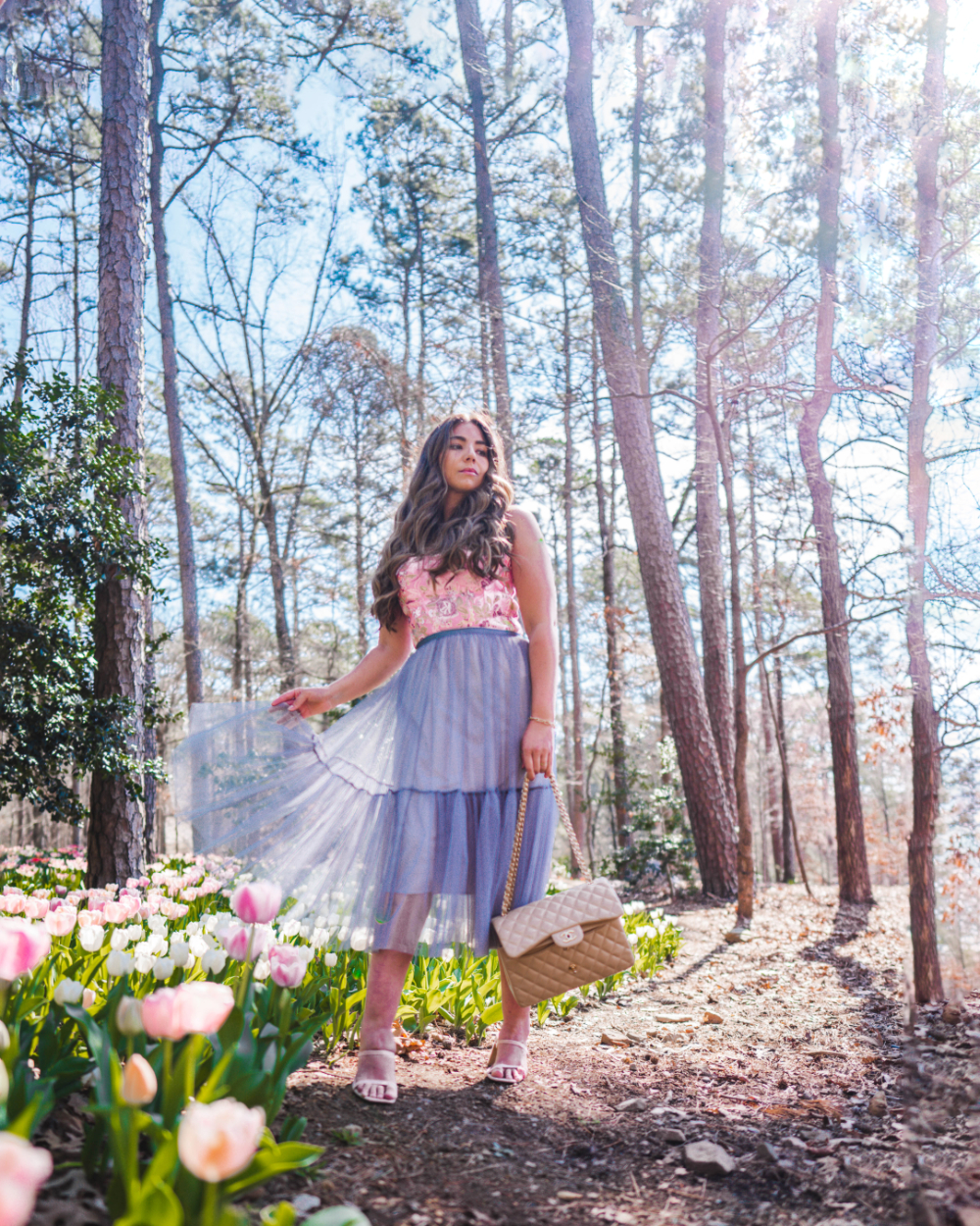 Fashion and style blogger Lauryncakes holds a tulle dress in one hand and a quilted purse in another while wearing a sleevless floral bustier and heels standing in the Garvan Gardens next to blooming tulips in the spring