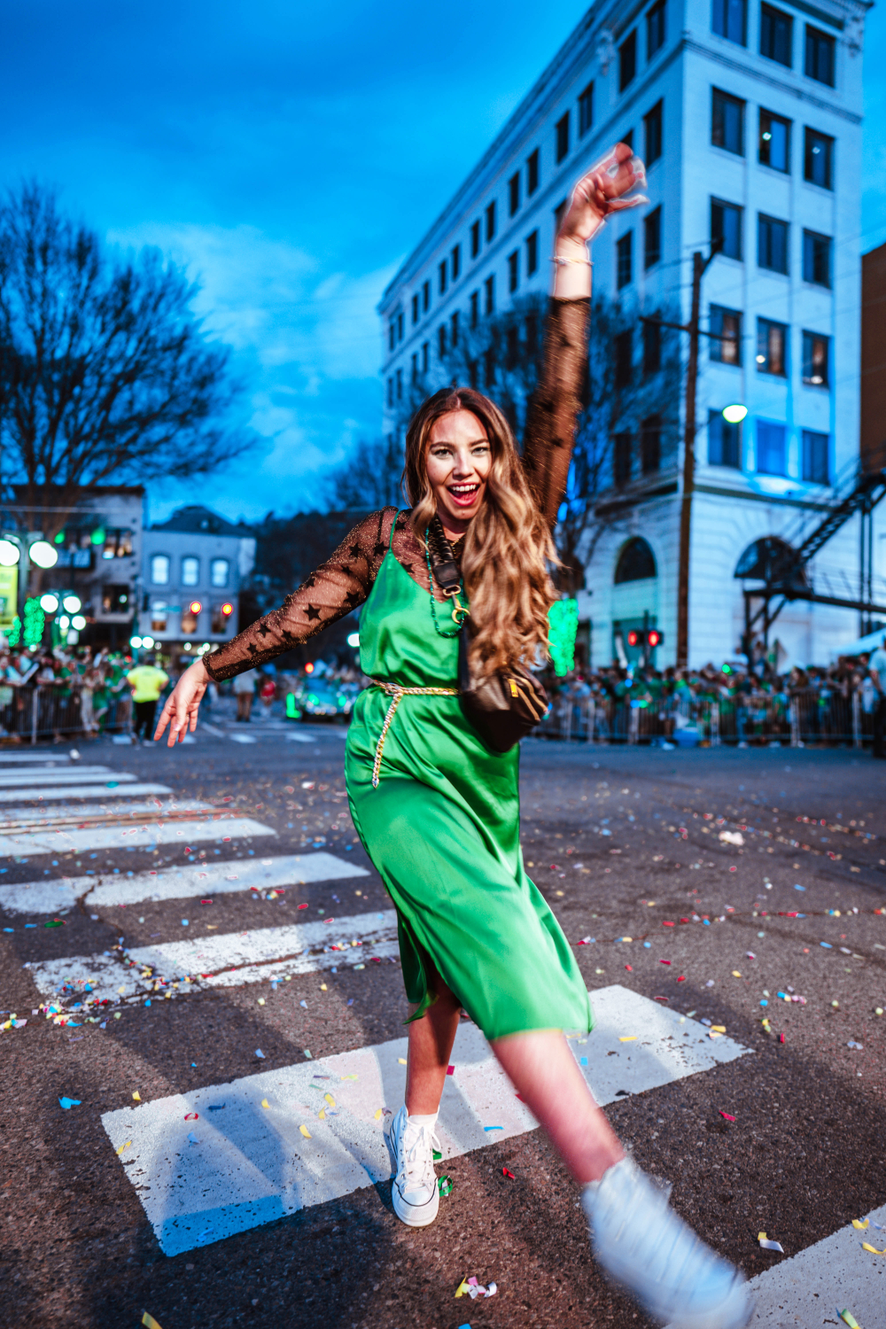 Lauryn Hock holds her hands in the air while wearing a star print mesh top, green satin slip dress, and belt at the Shortest St. Patricks Day Parade in Hot Springs Arkansas