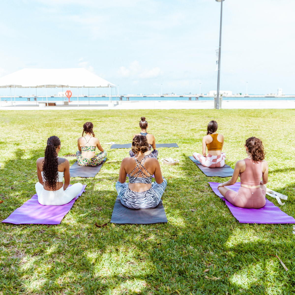 A group of women sit cross legged on yoga mats in the grass in Key West for a yoga session. All of the travel bloggers pictured are wearing workout outfits and facing the Atlantic ocean