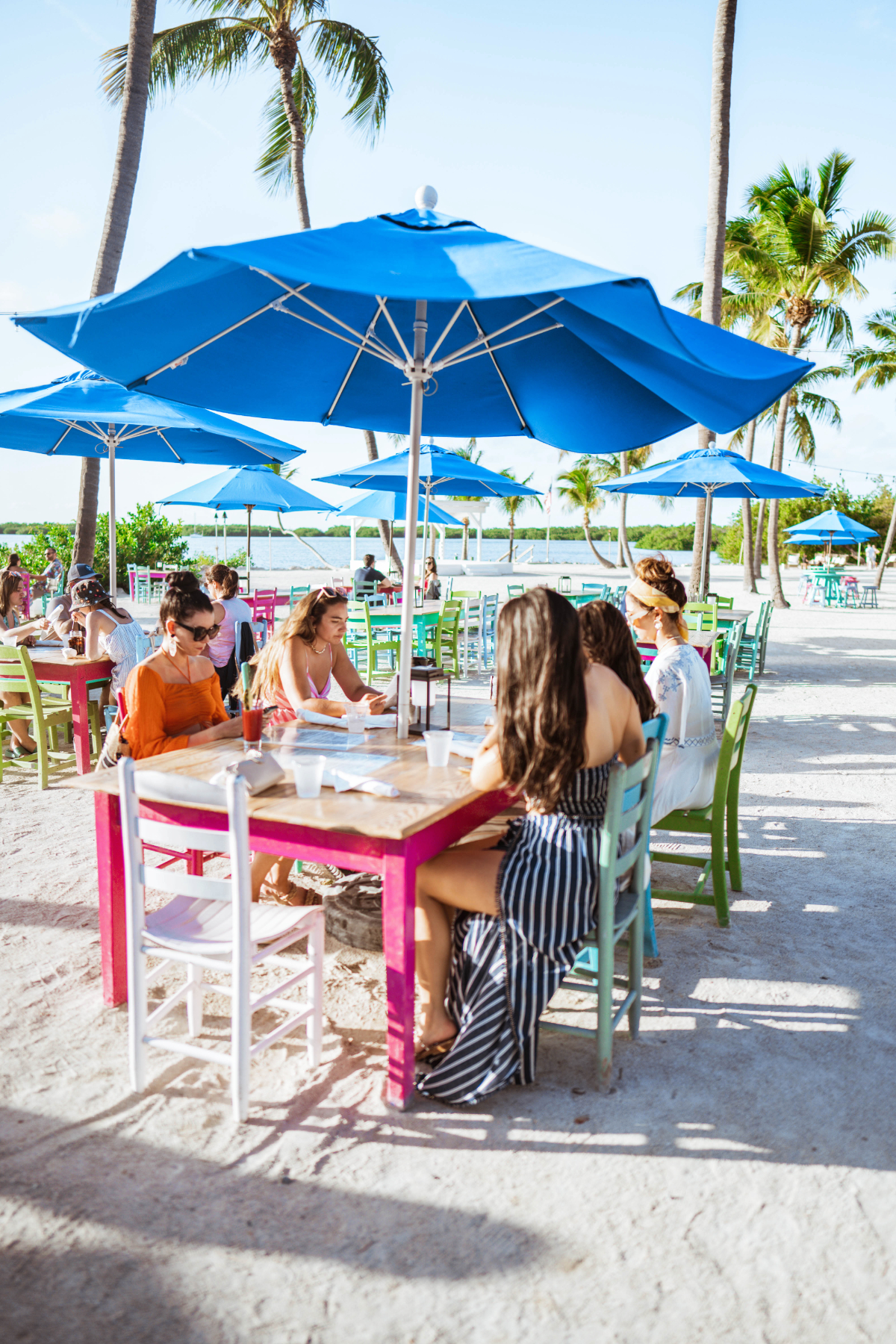 Five women sit at a wooden, colorful beach table under an umbrella looking at the dinner menu offered at Morada Bay Beach Cafe in Islamorada