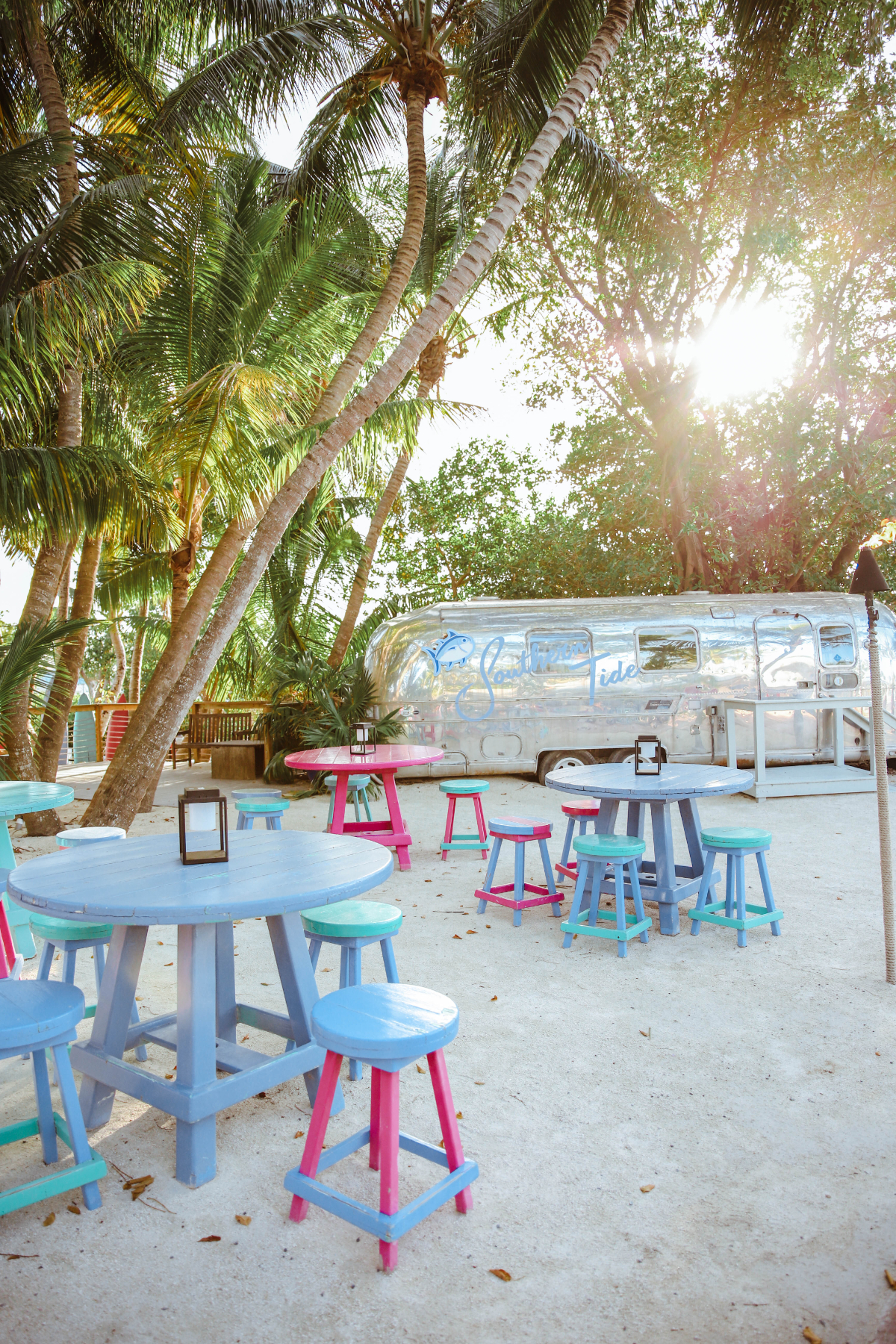 Wooden beach tables surround a metal food truck trailer and palm trees on a sandy area at Morada Bay Beach Cafe