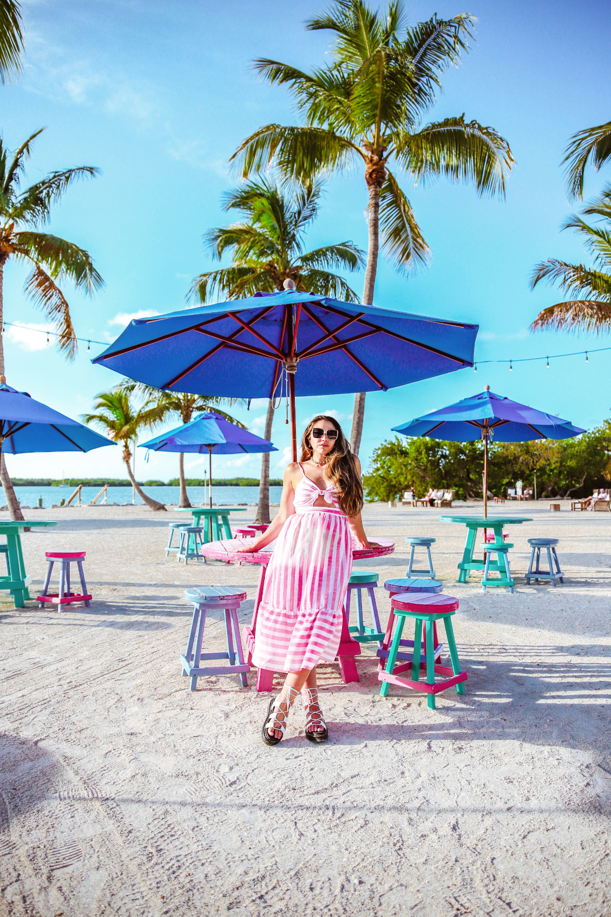 Travel blogger Lauryn Hock leans against a picnic table at Morada Bay Beach Cafe. She's wearing astriped pink maxi dress, sunglasses, and lace up sandals. Behind her are palm trees and other colorful beach tables, chairs, and umbrellas.