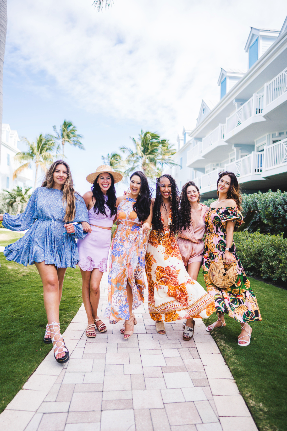 Group of six women wear island style dresses while linking arms and walking down a path in the middle of grass in Florida Keys